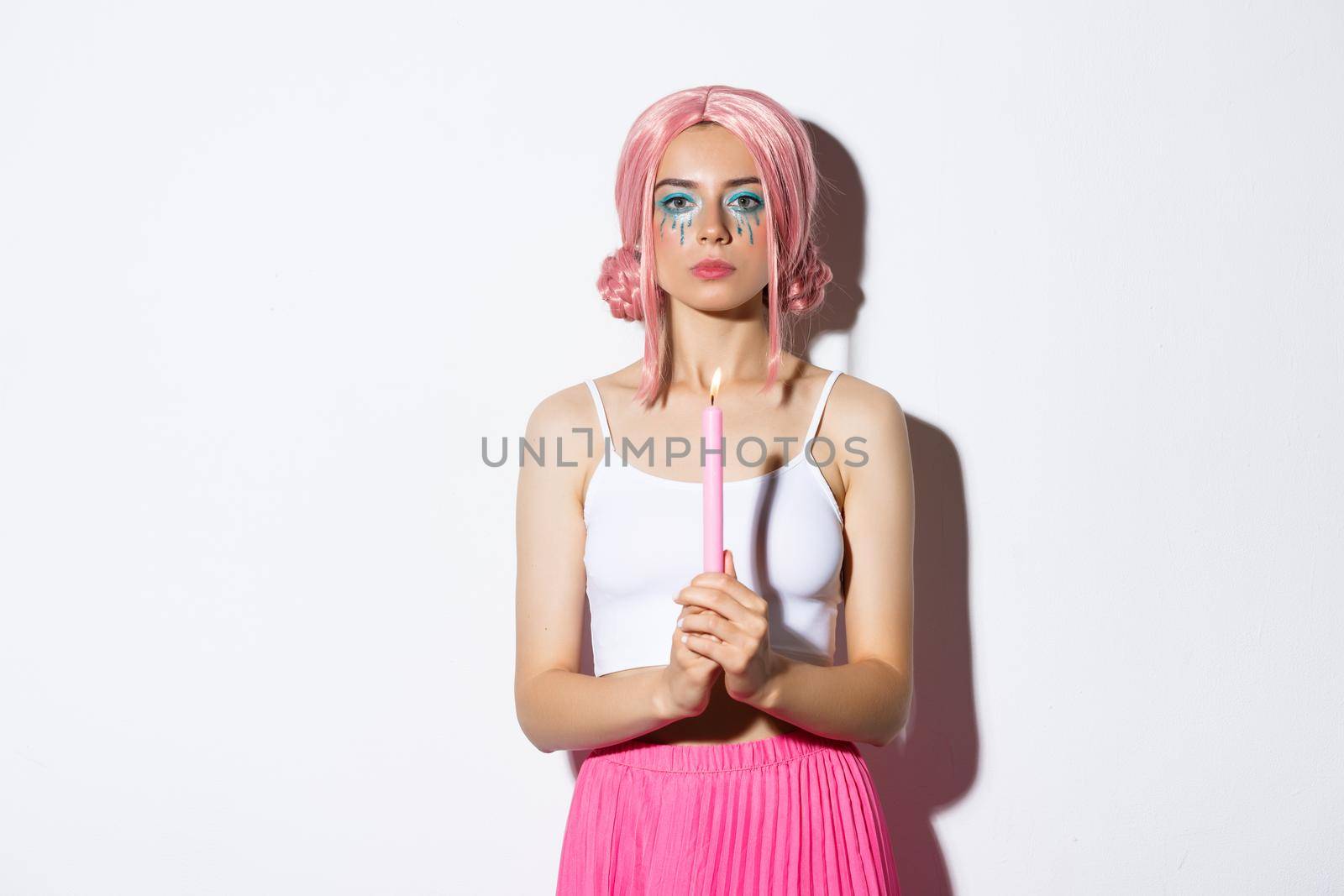 Image of serious looking girl dressed-up as fairy on halloween, holding candle, standing over white background in pink wig.