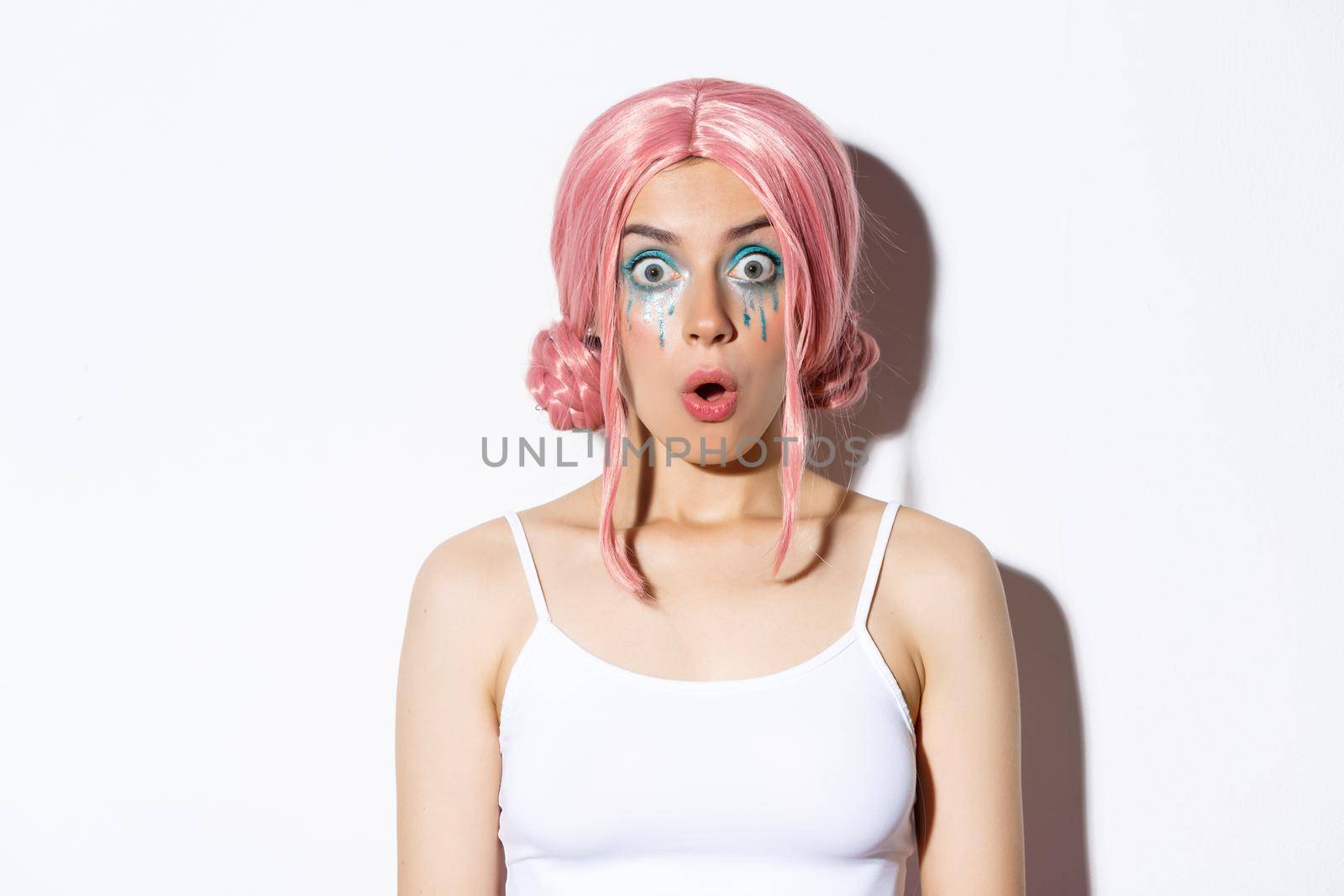 Close-up of surprised pretty girl in pink wig, gasping and looking amazed, standing over white background.