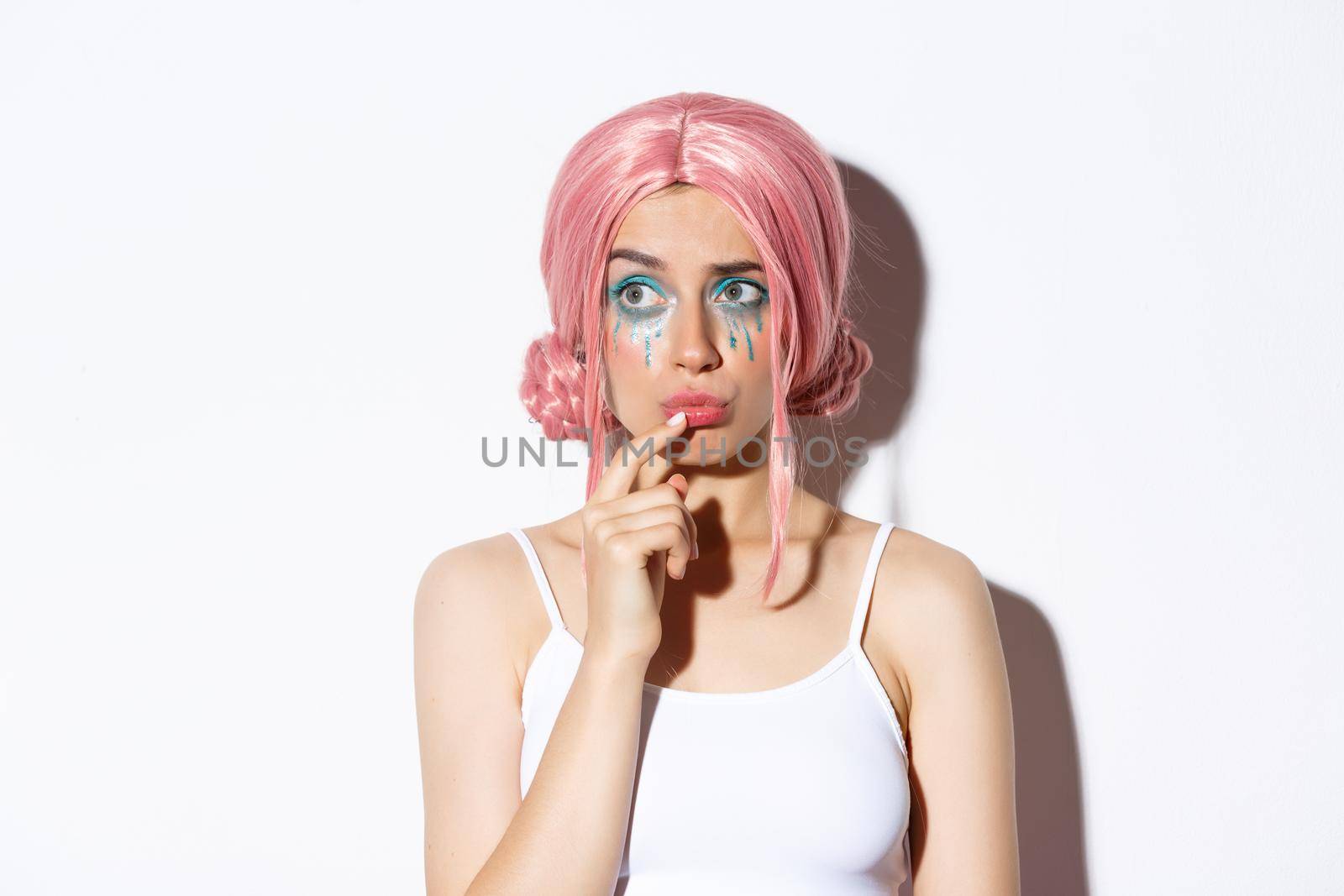 Close-up of concerned cute girl in pink party wig, looking left and thinking, standing over white background in halloween costume.