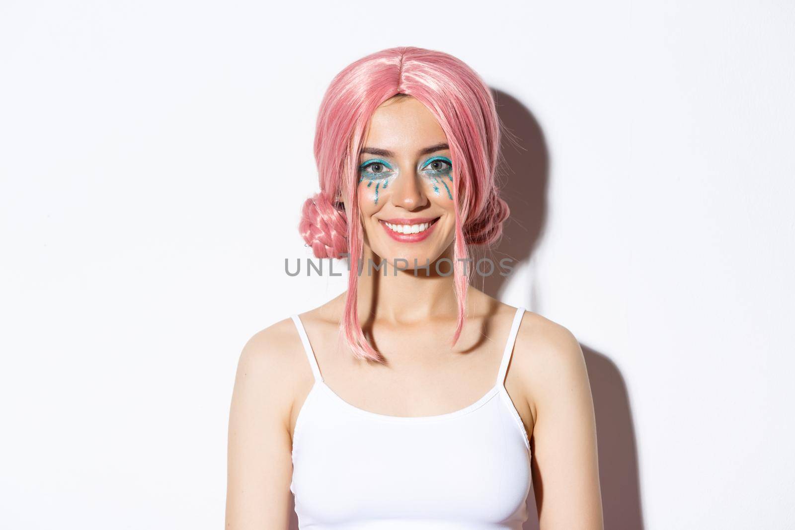 Close-up of pretty female model in pink wig, colorful makeup, dressed up for halloween party, standing over white background.