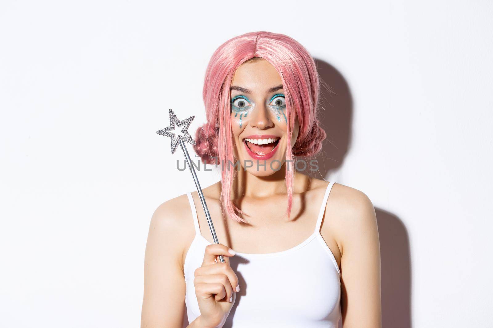 Close-up of surprised happy girl in halloween costume of fairy, holding magic wand and smiling amazed at camera, standing over white background.