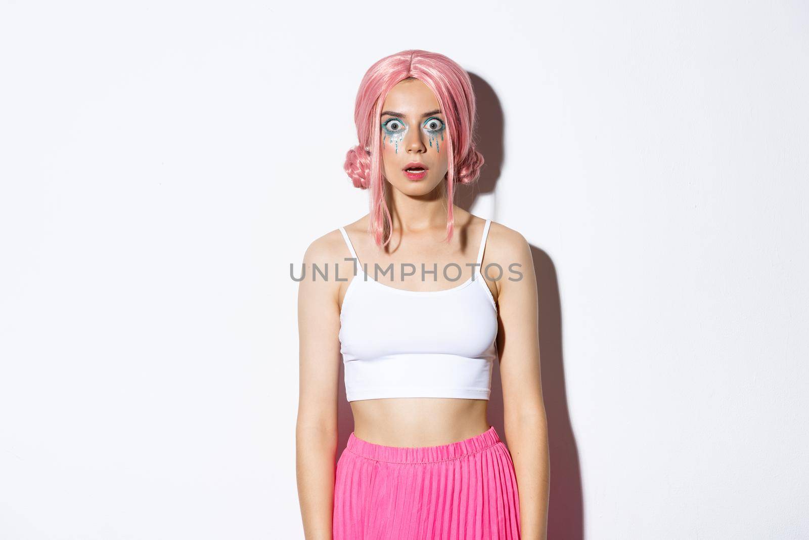 Portrait of glamorous party girl in pink wig and bright makeup, drop jaw and looking startled at camera, stare at something impressive, standing over white background.