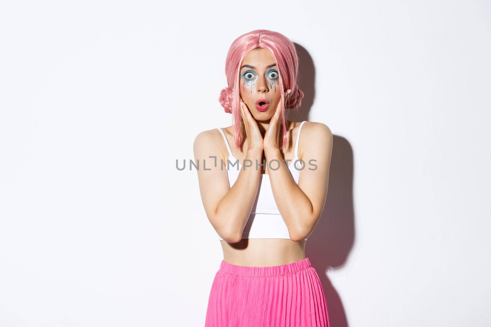 Portrait of surprised caucasian girl in pink wig and bright makeup, gasping amazed and stare at camera in awe, standing over white background in halloween party costume.