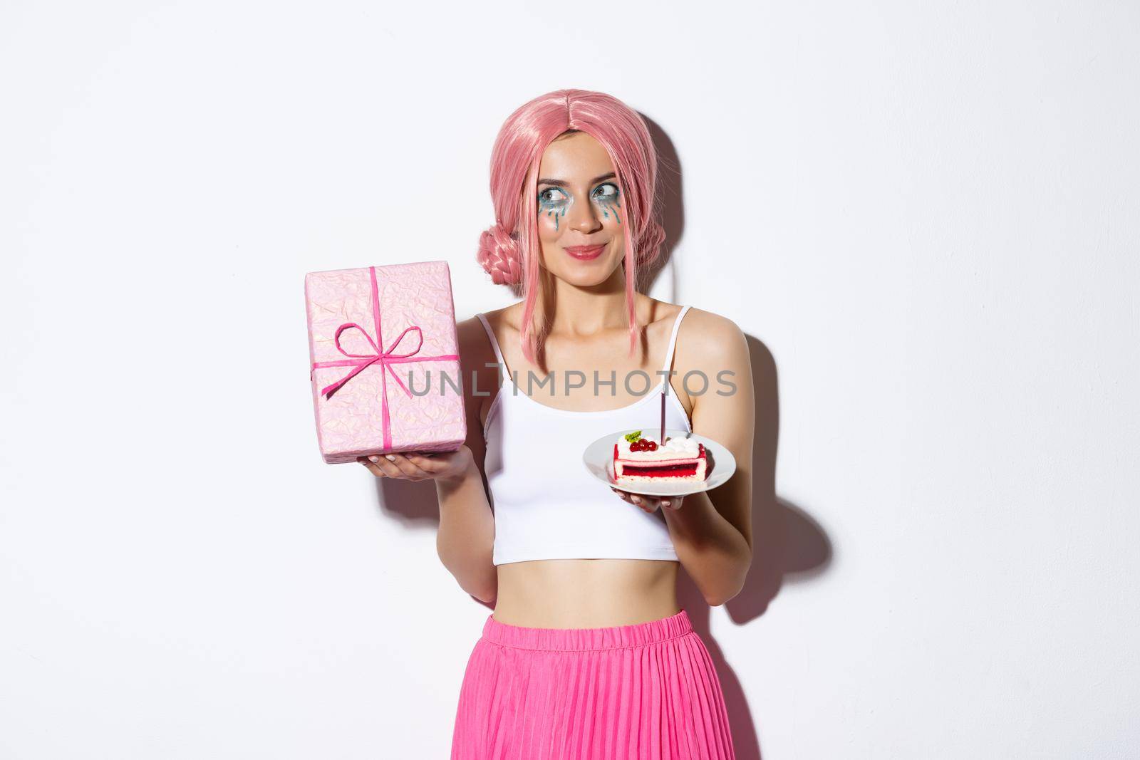 Image of excited cute girl in pink wig, shaking box with gift and wander what inside, holding piece of birthday cake, celebrating b-day.