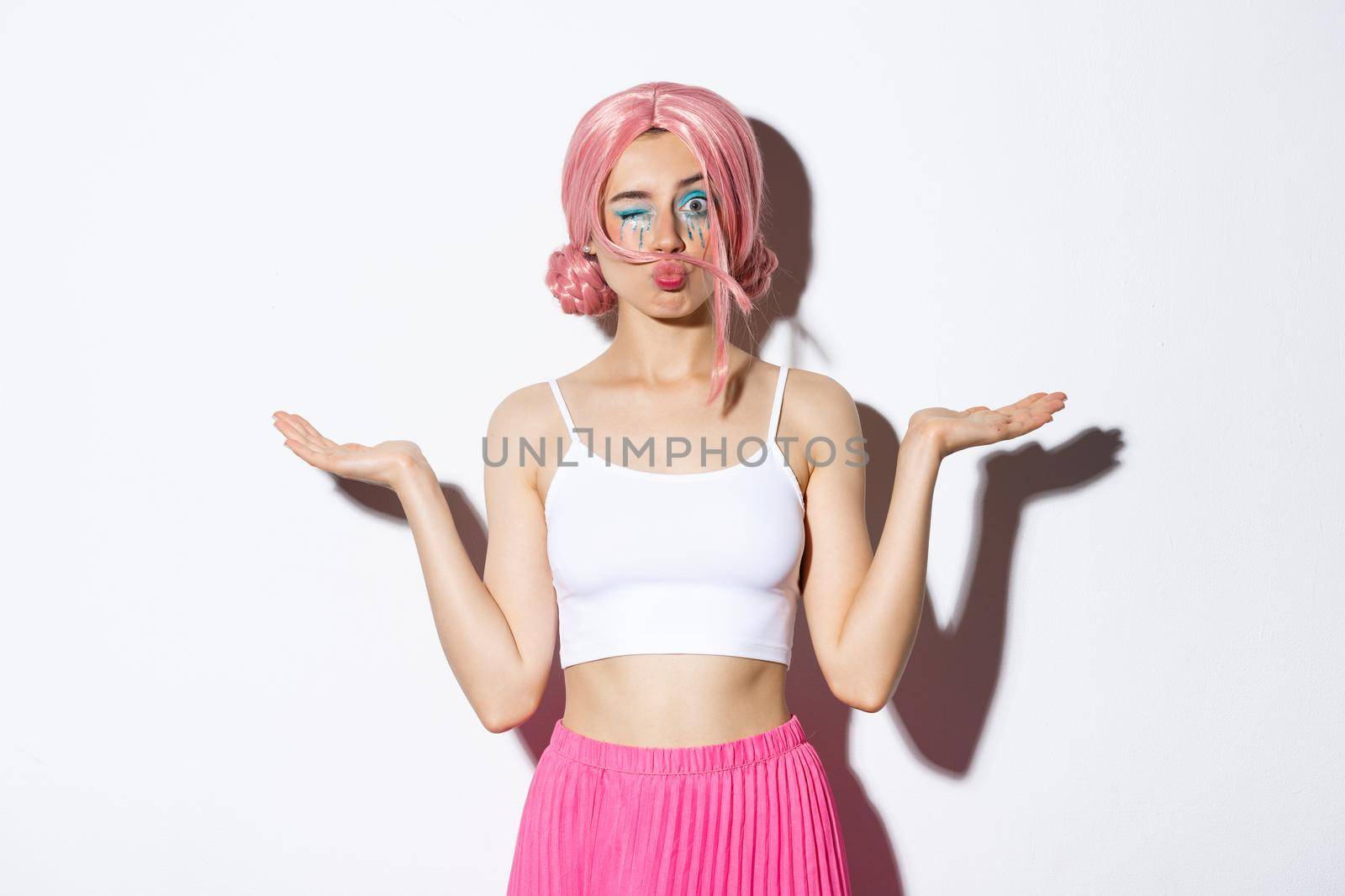Portrait of silly pop girl wearing pink wig, winking at camera while holding something in both hands, standing over white background.