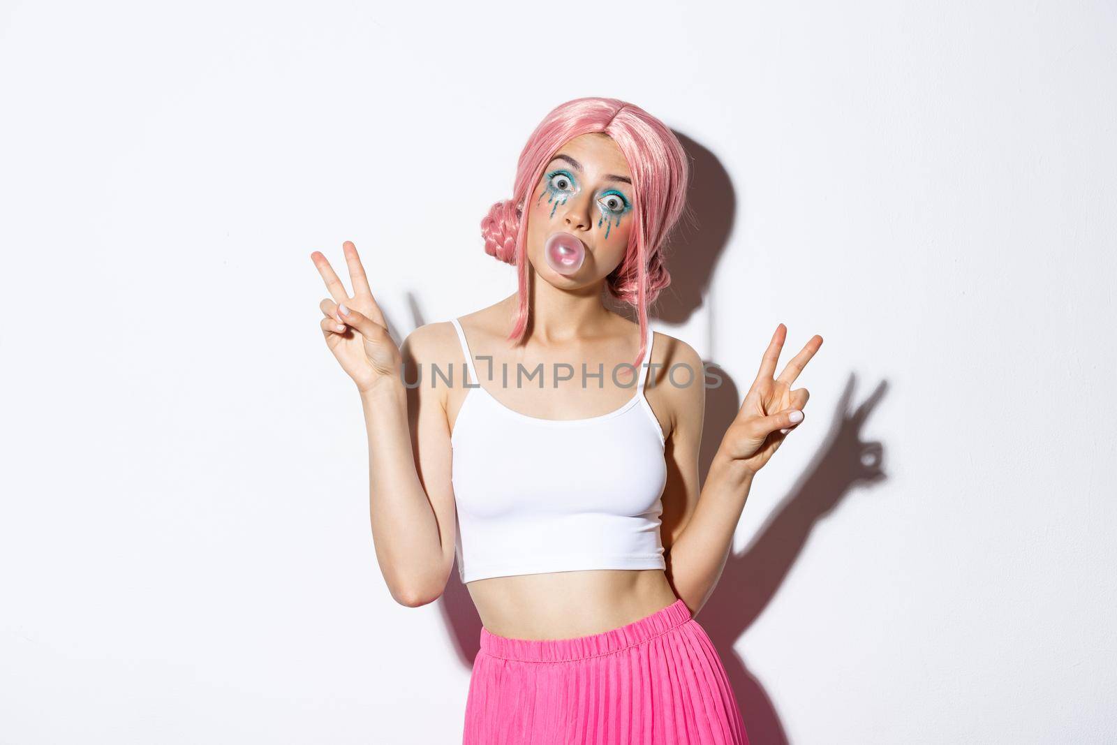 Image of beautiful girl in pink wig blowing chewing gum, popping bubble on face and showing peace signs, standing silly over white background.