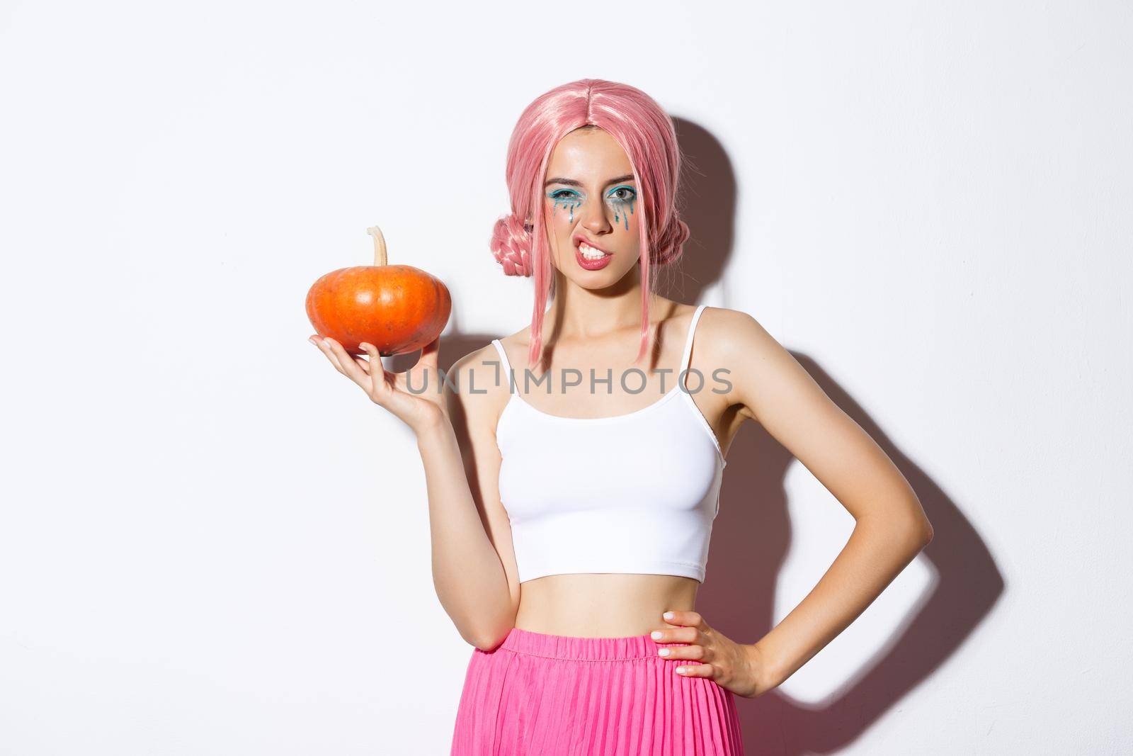 Portrait of sassy young female model in pink wig, holding pumpkin, celebrating halloween, standing over white background.