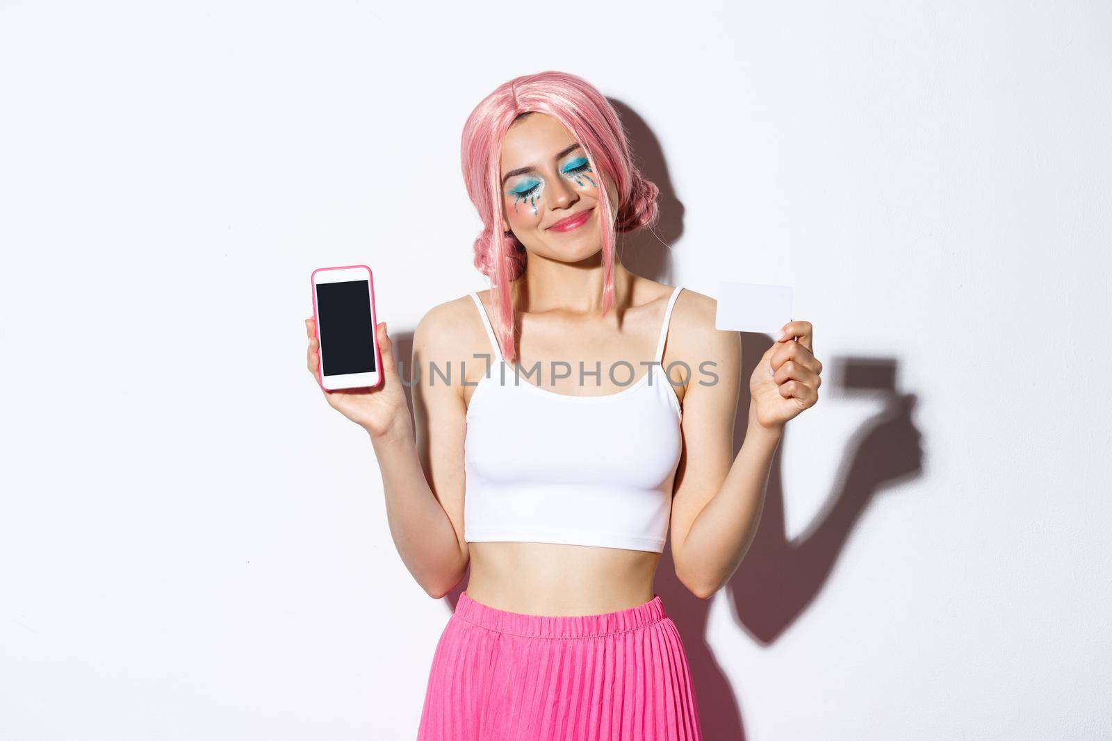 Portrait of lovely pleased girl looking happy, close eyes and smiling, showing credit card with smartphone screen, standing in pink anime wig and bright makeup.
