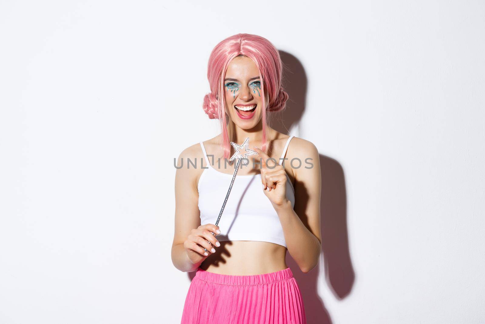 Portrait of attractive girl with pink wig and bright makeup, dressed up as a fairy for halloween party, holding magic wand and smiling.