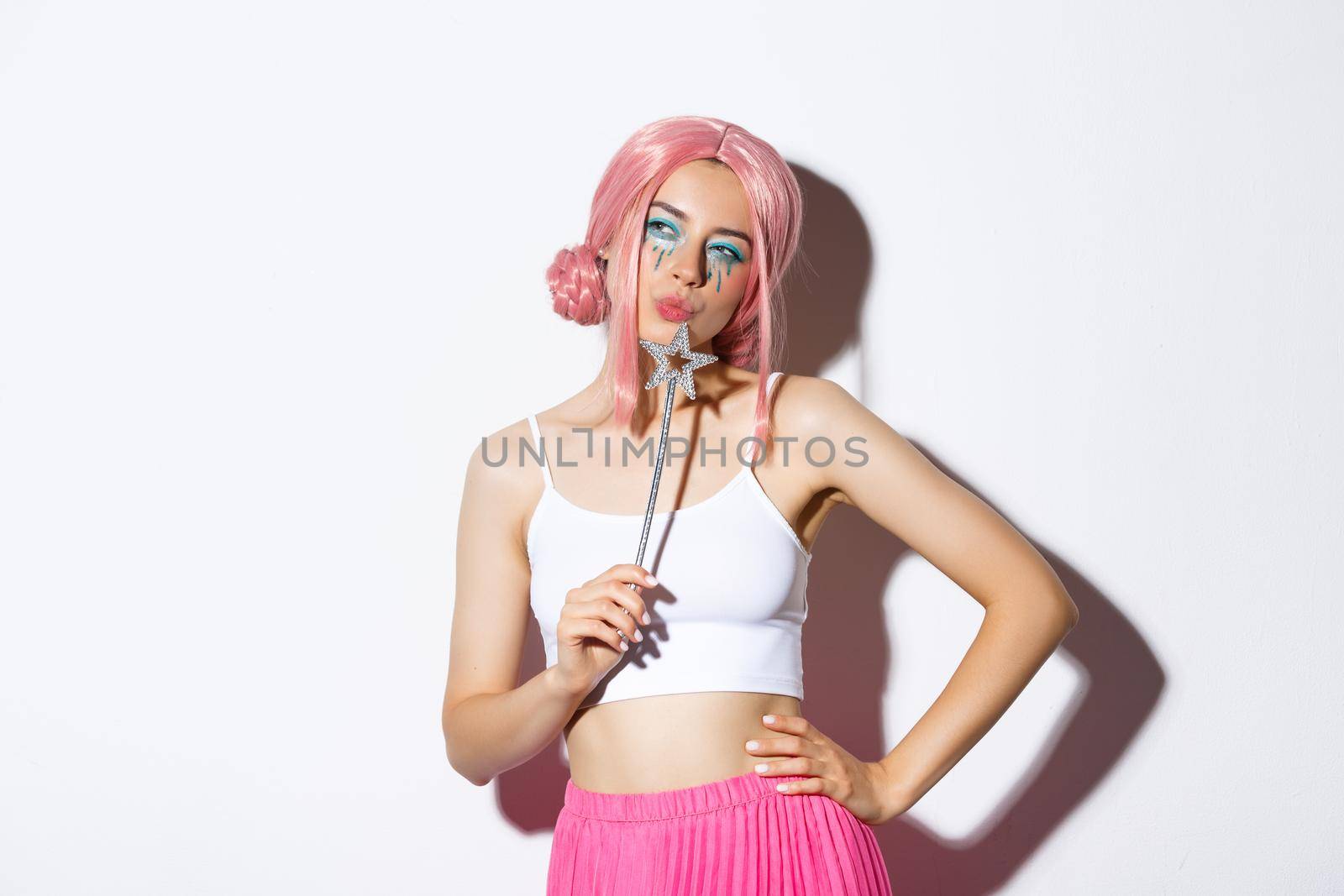 Portrait of beautiful girl in fairy costume looking thoughtful, holding magic wand and wearing pink wig, standing over white background, celebrating halloween.