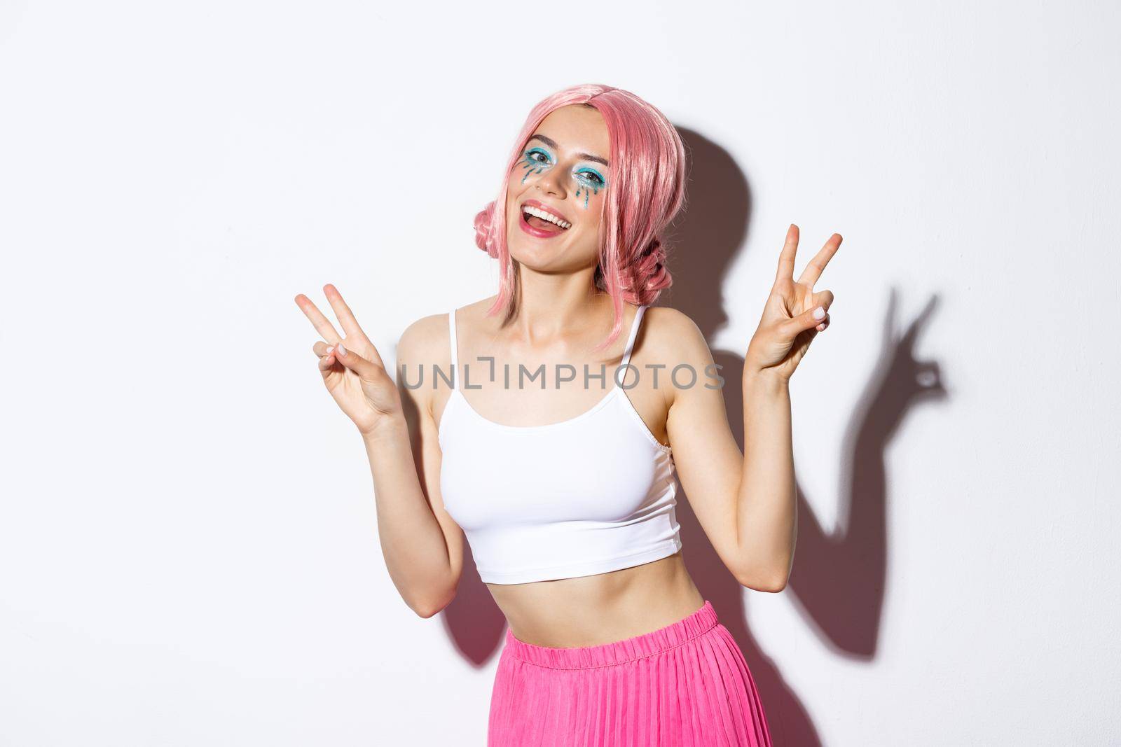 Portrait of beautiful smiling girl in pink wig, having fun on party, showing peace signs, celebrating holiday, standing over white background.