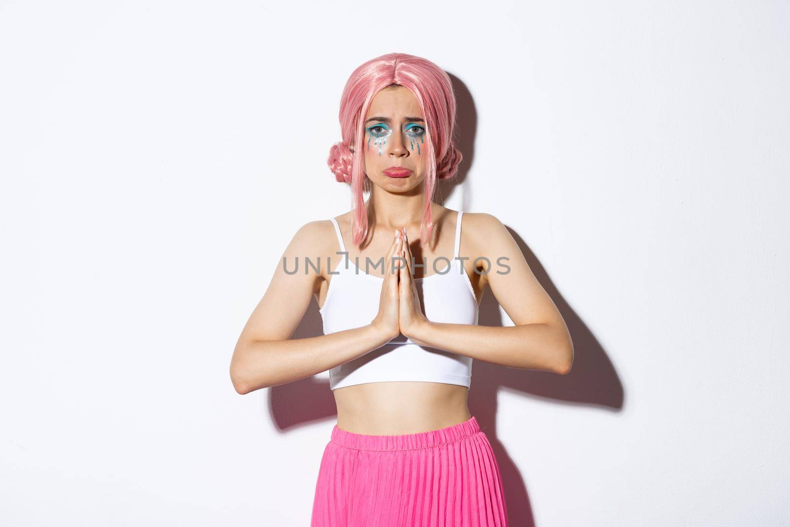 Portrait of miserable cute girl in pink anime wig asking for help, clasp hands together and begging for something, standing in halloween outfit over white background.