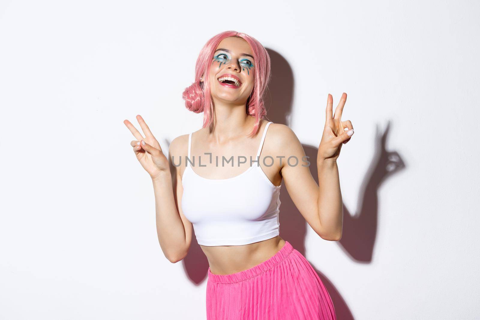 Portrait of beautiful smiling girl in pink wig, showing kawaii peace signs and laughing, wearing outfit for party, standing over white background.