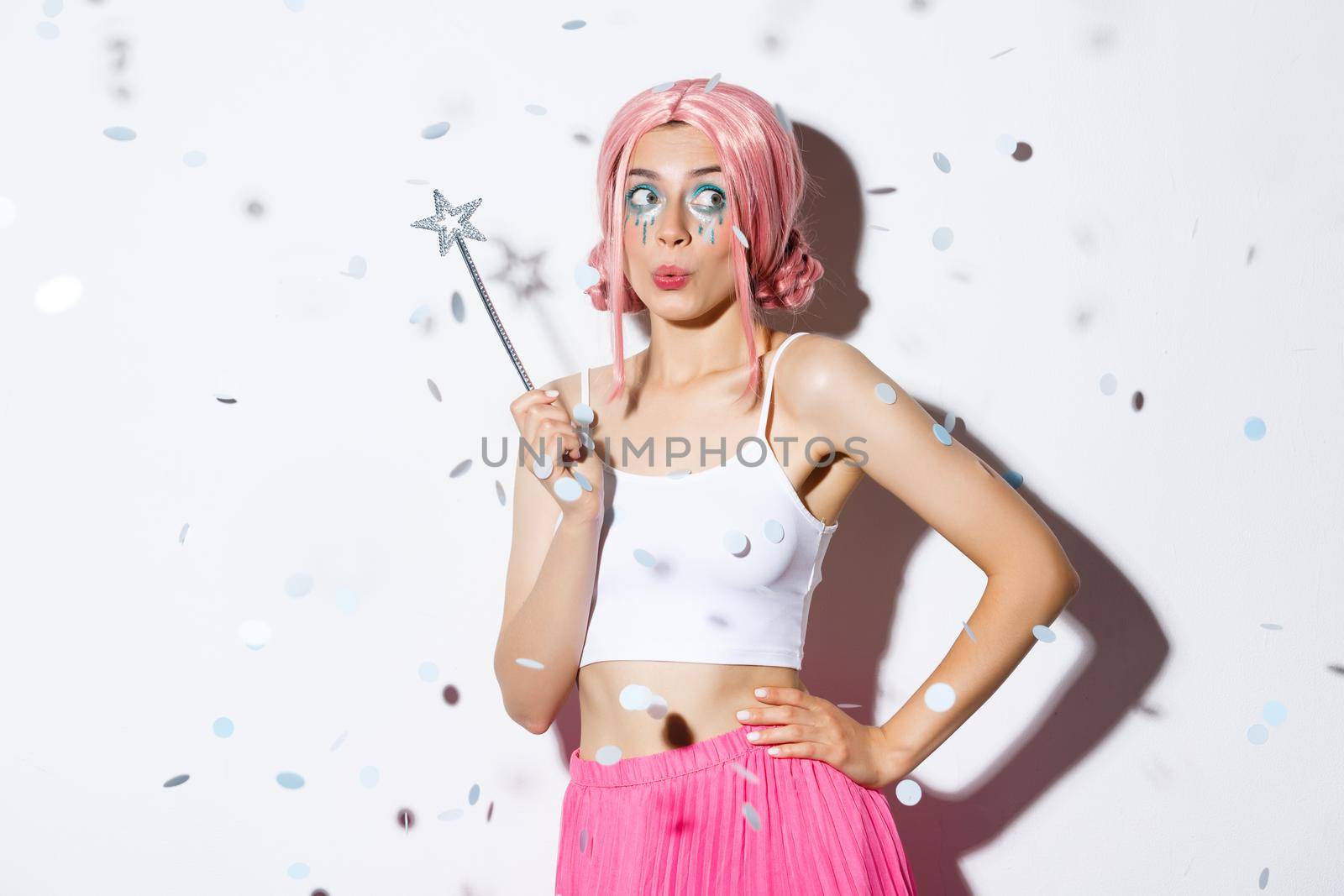 Image of beautiful happy girl in pink wig, celebrating halloween in fairy costume, holding magic wand and smiling while confetti floating in air.