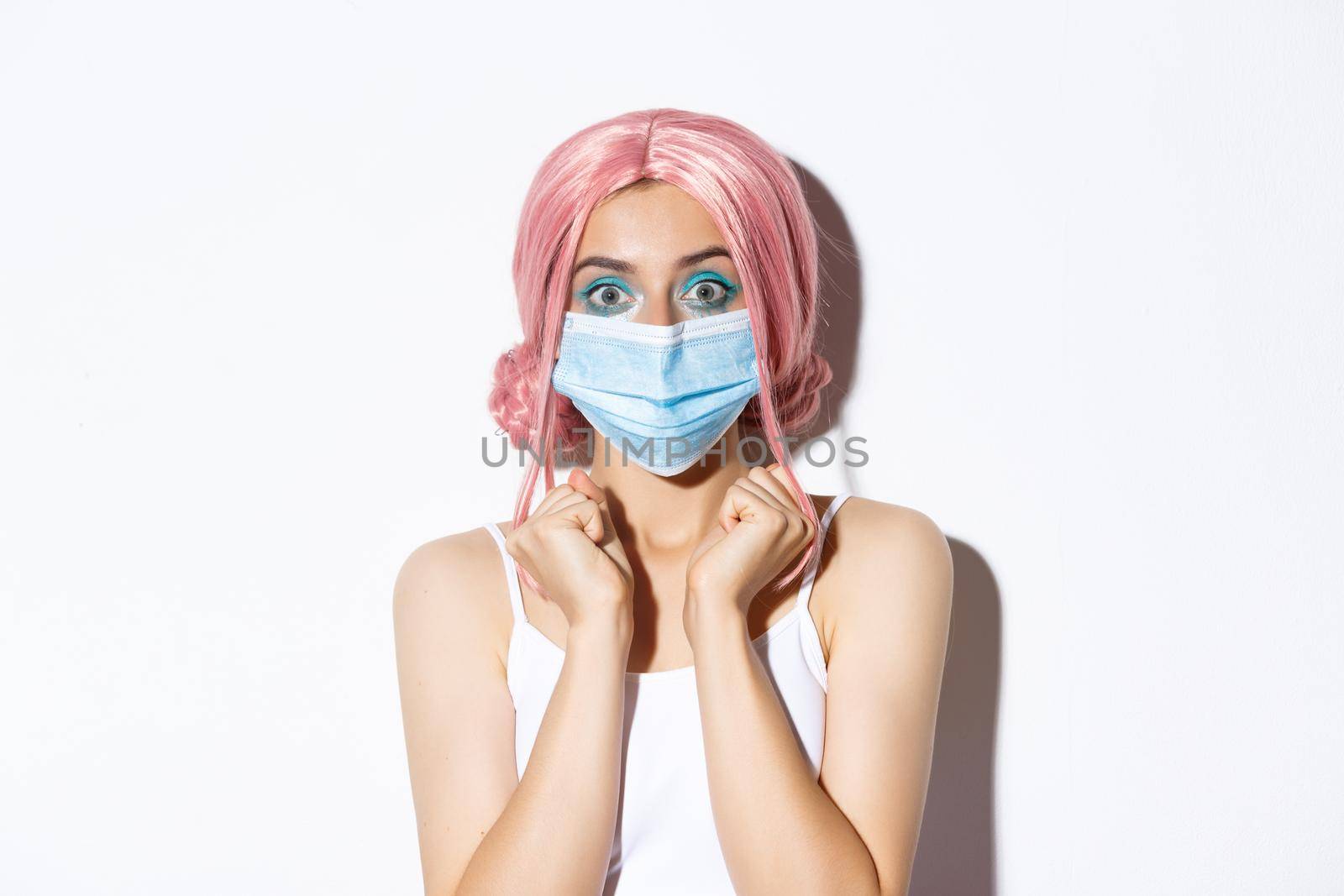 Coronavirus, social distancing and lifestyle concept. Close-up of excited attractive girl in pink wig and medical mask, looking with hope at camera, standing over white background.