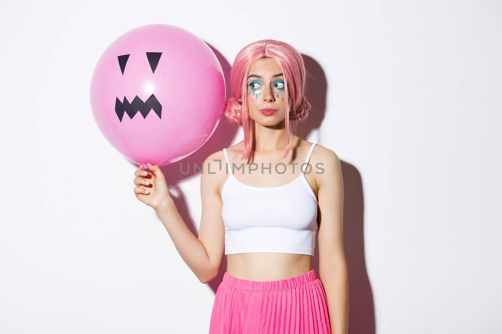 Image of beautiful glamour girl in pink wig, bright makeup, holding balloon with scary halloween face, standing over white bacground.
