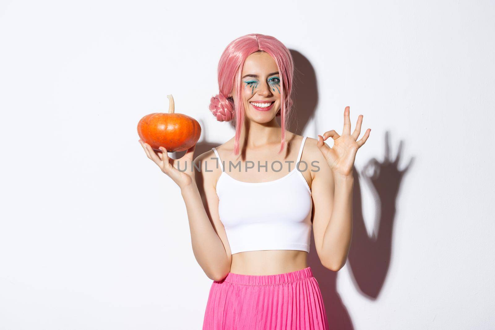 Image of happy girl celebrating halloween, wearing pink wig, holding pumpkin and showing okay sign, standing over white background by Benzoix
