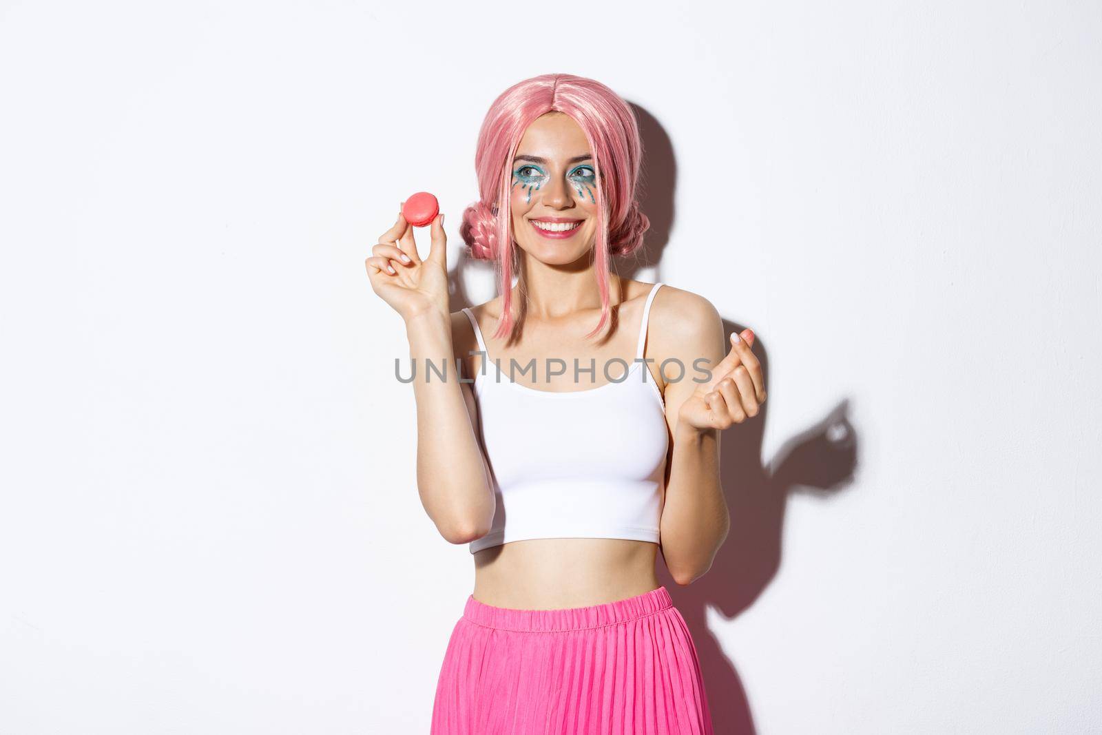 Portrait of lovely female model eating macaroons and smiling, wearing pink wig and outfit for party, standing over white background by Benzoix