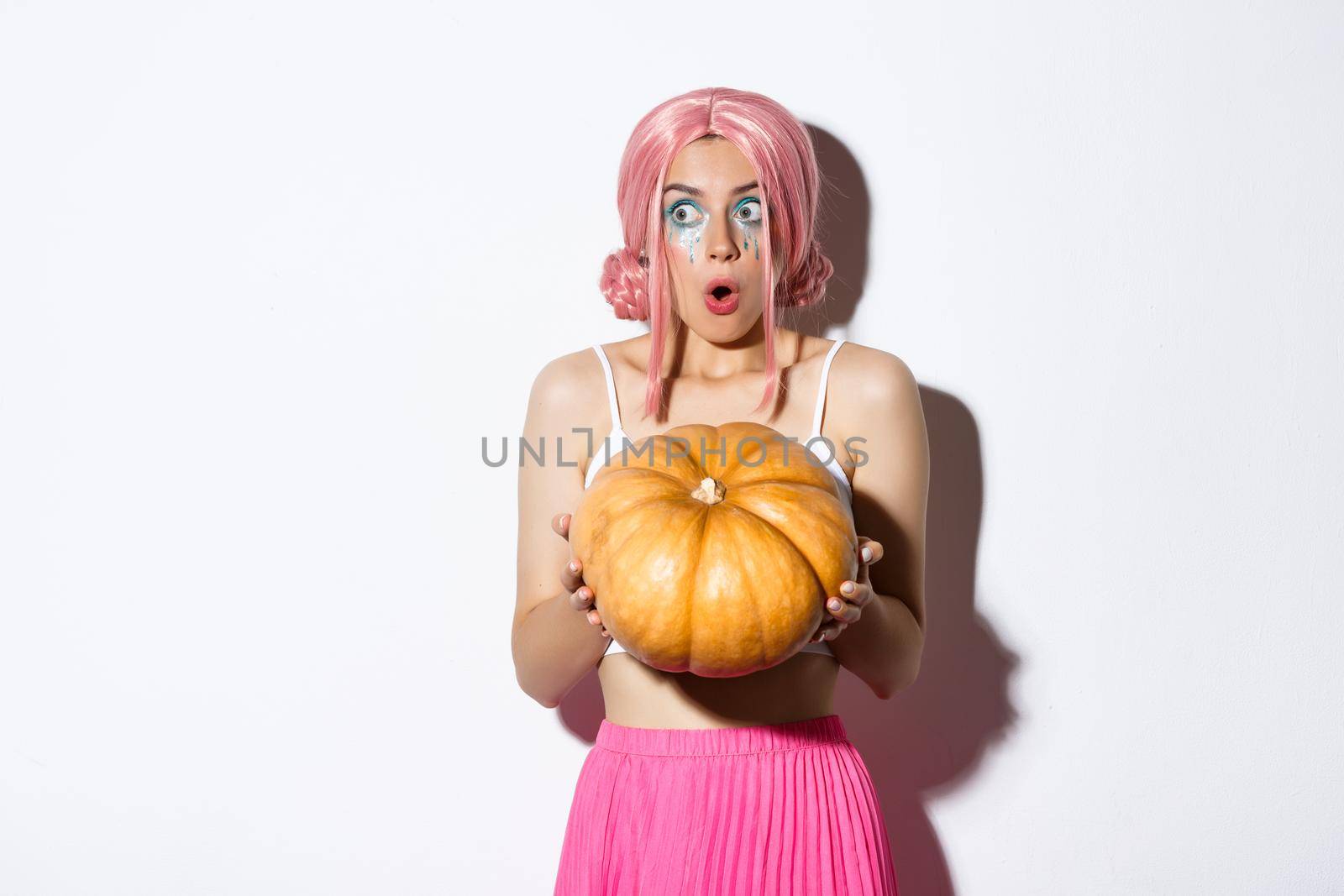 Portrait of amazed young party girl in pink wig, holding big pumpkin and looking left in awe, celebrating halloween, standing over white background.