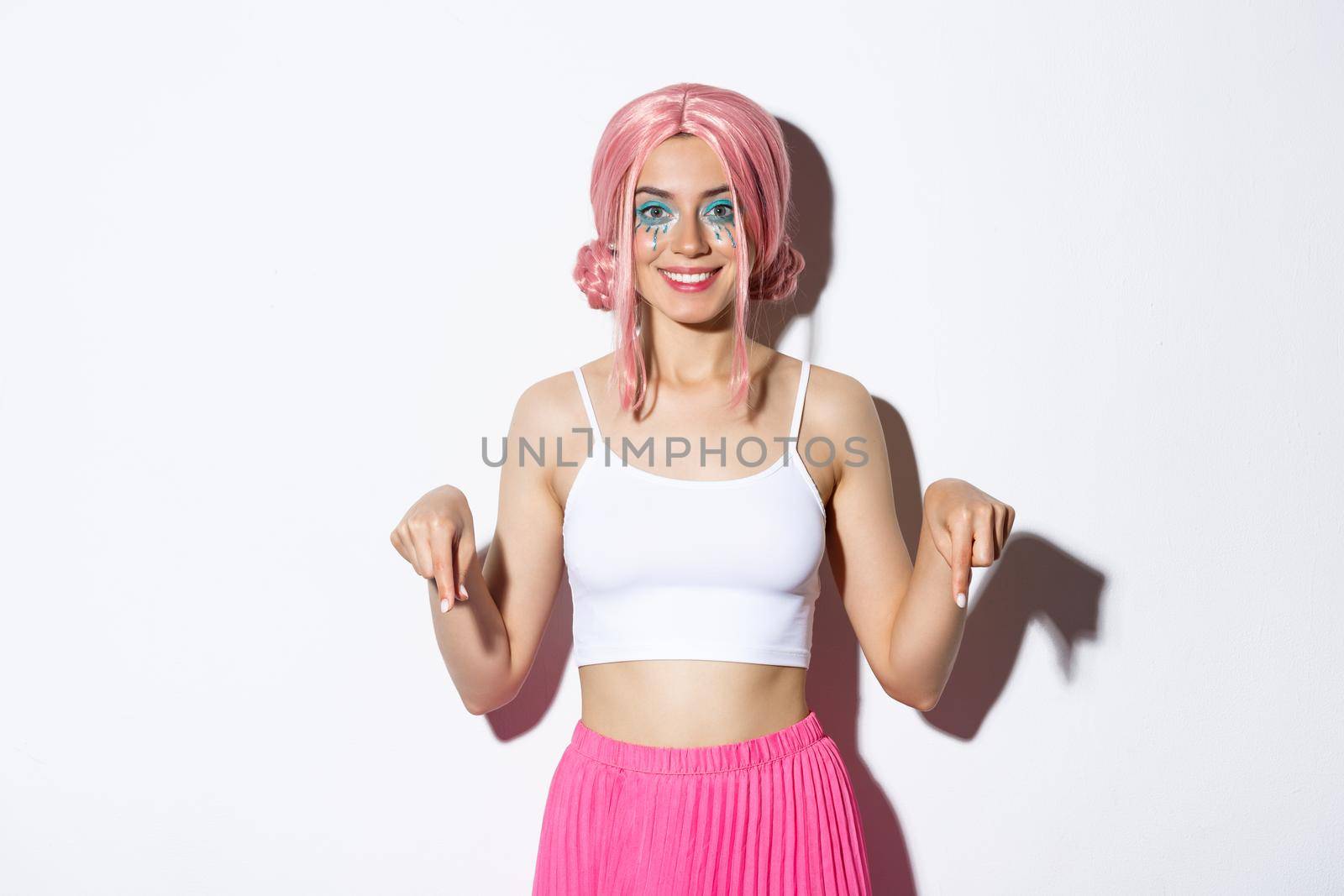 Beautiful smiling party girl with pink hair and bright makeup, pointing fingers down at your logo, wearing halloween costume, standing over white background by Benzoix