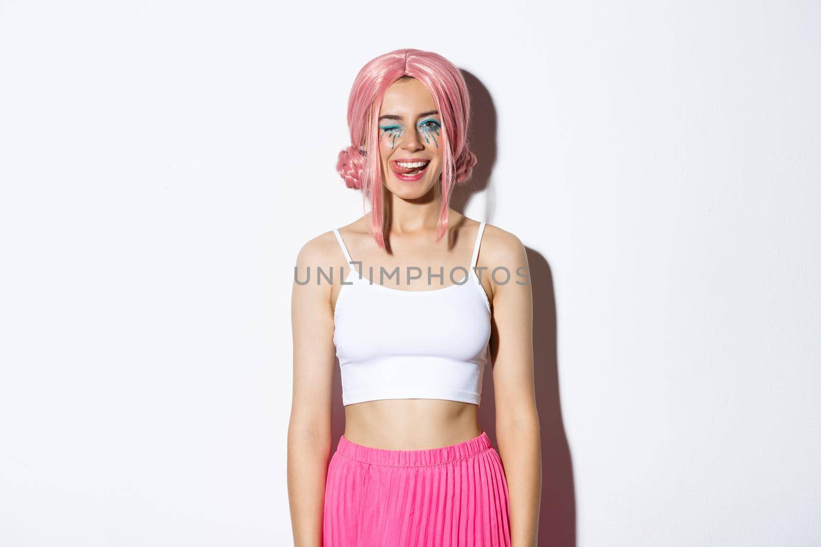 Portrait of carefree smiling female model in pink party wig, winking and showing tongue, celebrating halloween, standing over white background by Benzoix
