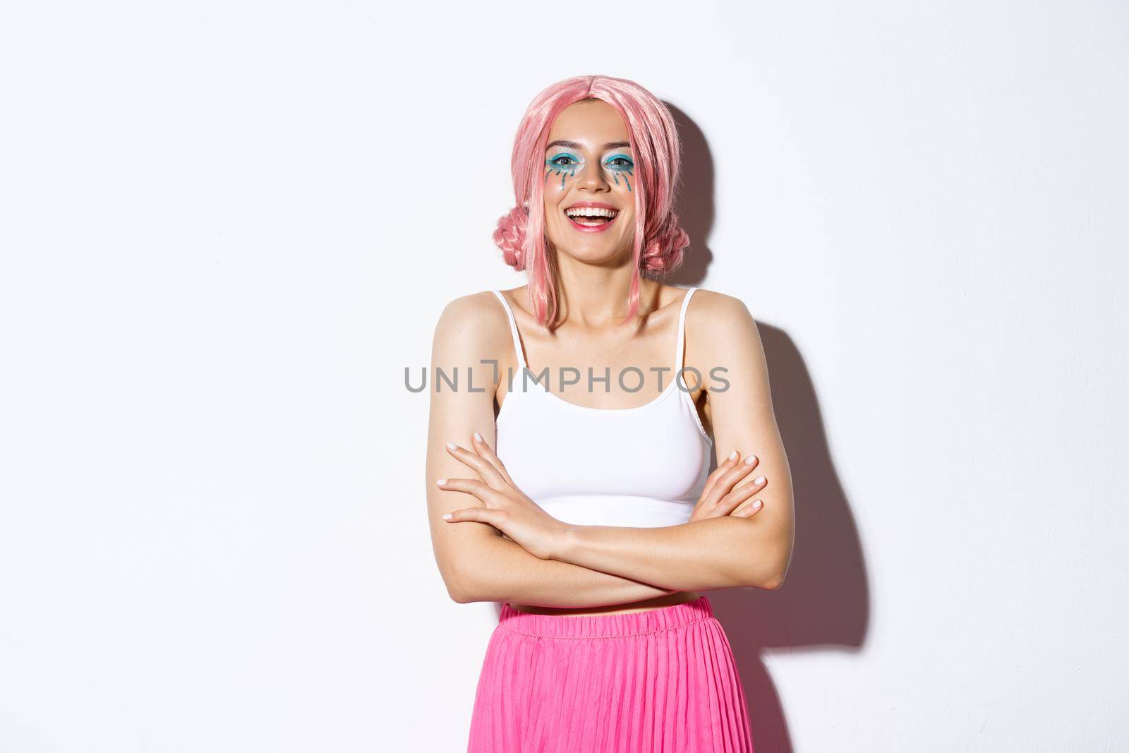 Indoor shot of carefree female model in pink wig, colorful makeup, dressed up for halloween party, standing over white background with arms crossed and confident smile.