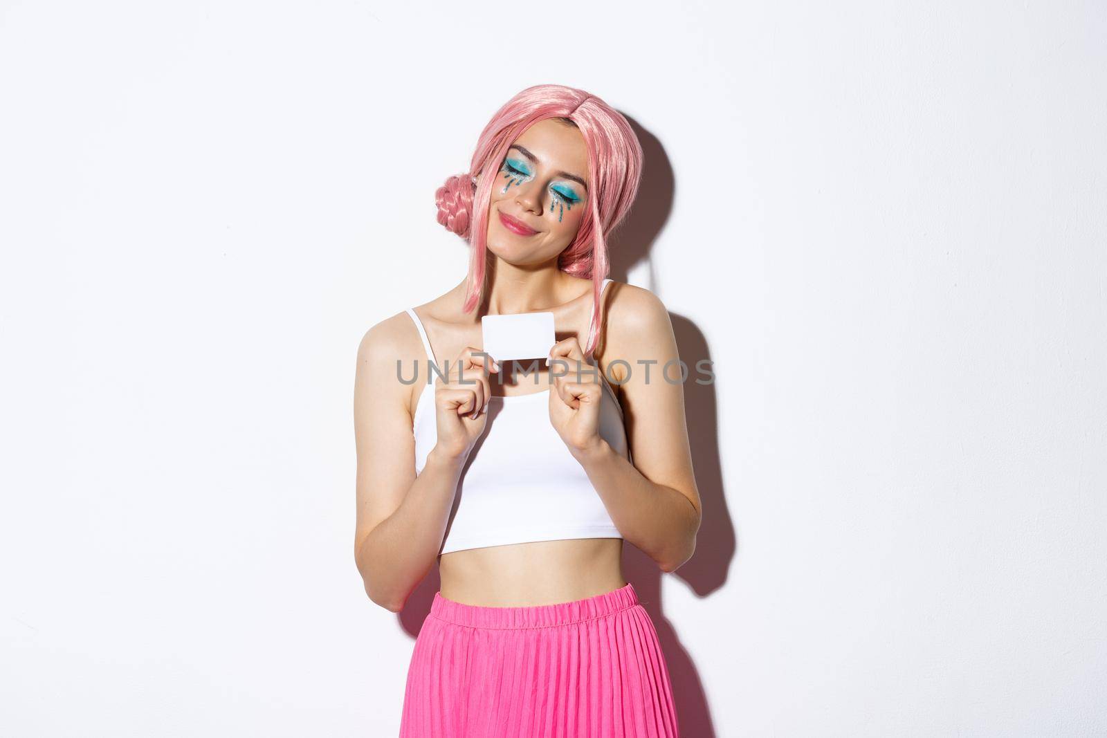 Portrait of attractive girl in pink wig, dreaming about shopping, holding credit card and smiling pleased, standing over white background.
