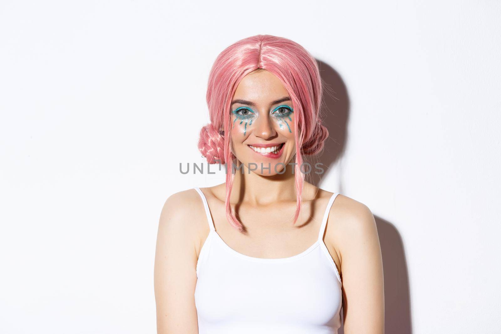 Close-up of flirty attractive female model in pink wig, with bright party makeup, biting lip and smiling, looking at something tempting, standing over white background.