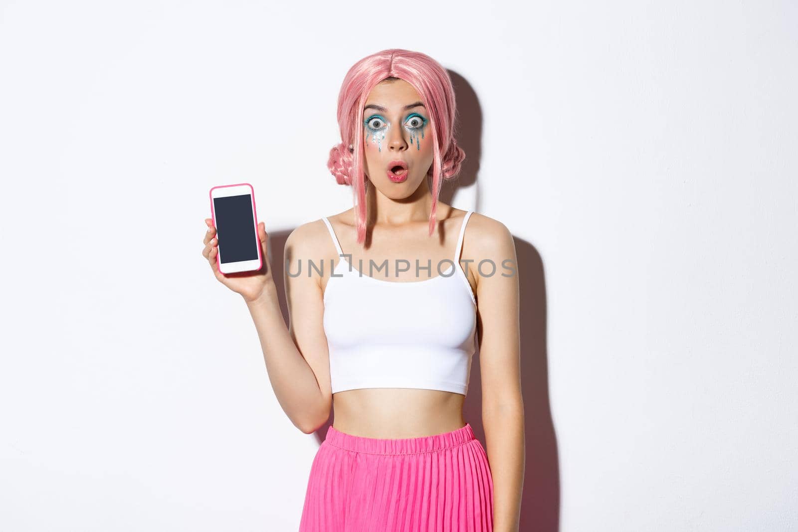 Image of surprised girl looking in awe while showing smartphone screen, dressed in pink wig and party outfit, standing over white background.