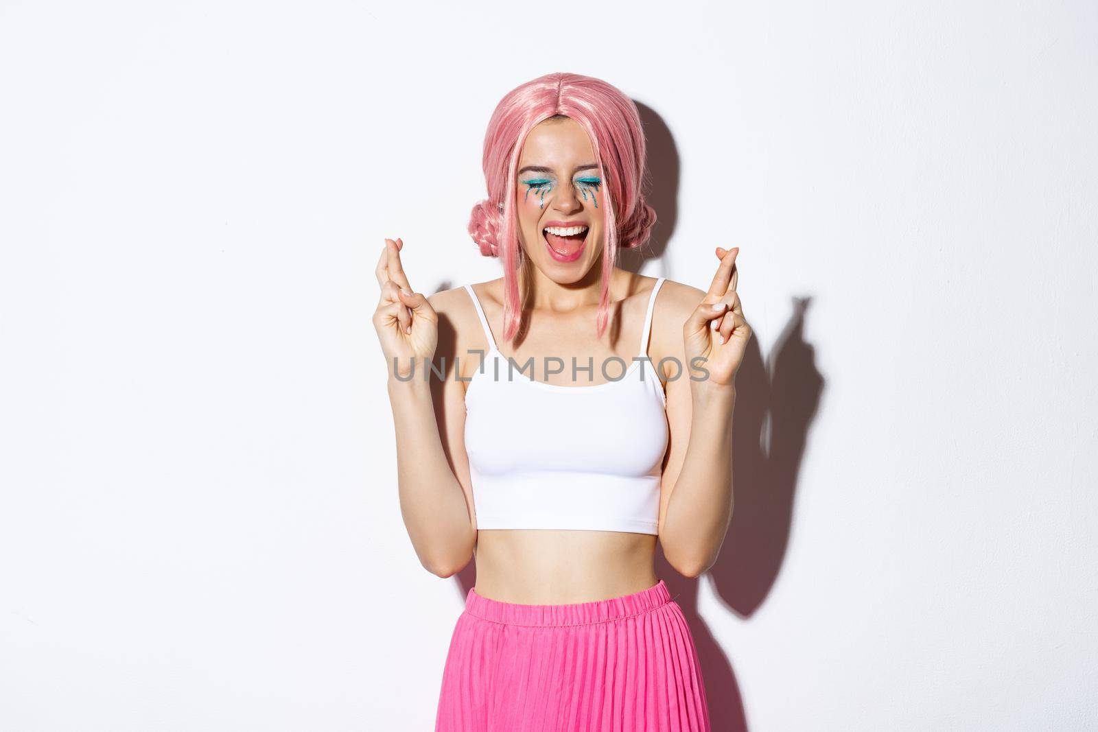 Image of optimistic party girl in pink wig, with halloween makeup, close eyes and screaming excited while cross fingers to make a wish, standing over white background by Benzoix