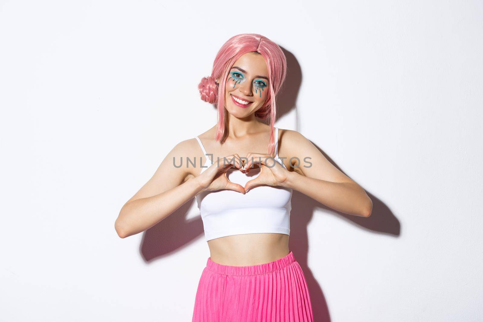Portrait of lovely girl in pink party wig and bright makeup, showing heart gesture and smiling, standing over white background.