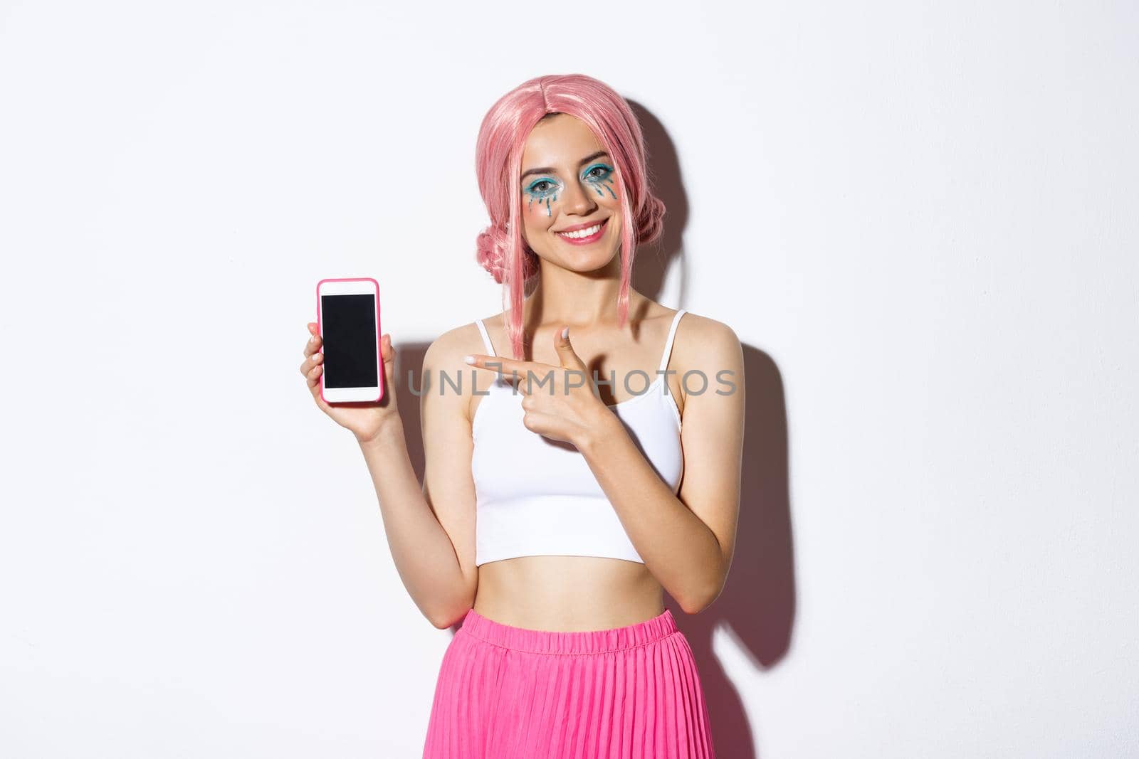 Portrait of happy beautiful female model in pink glamour wig and bright makeup, pointing finger at mobile phone screen, showing application or banner by Benzoix