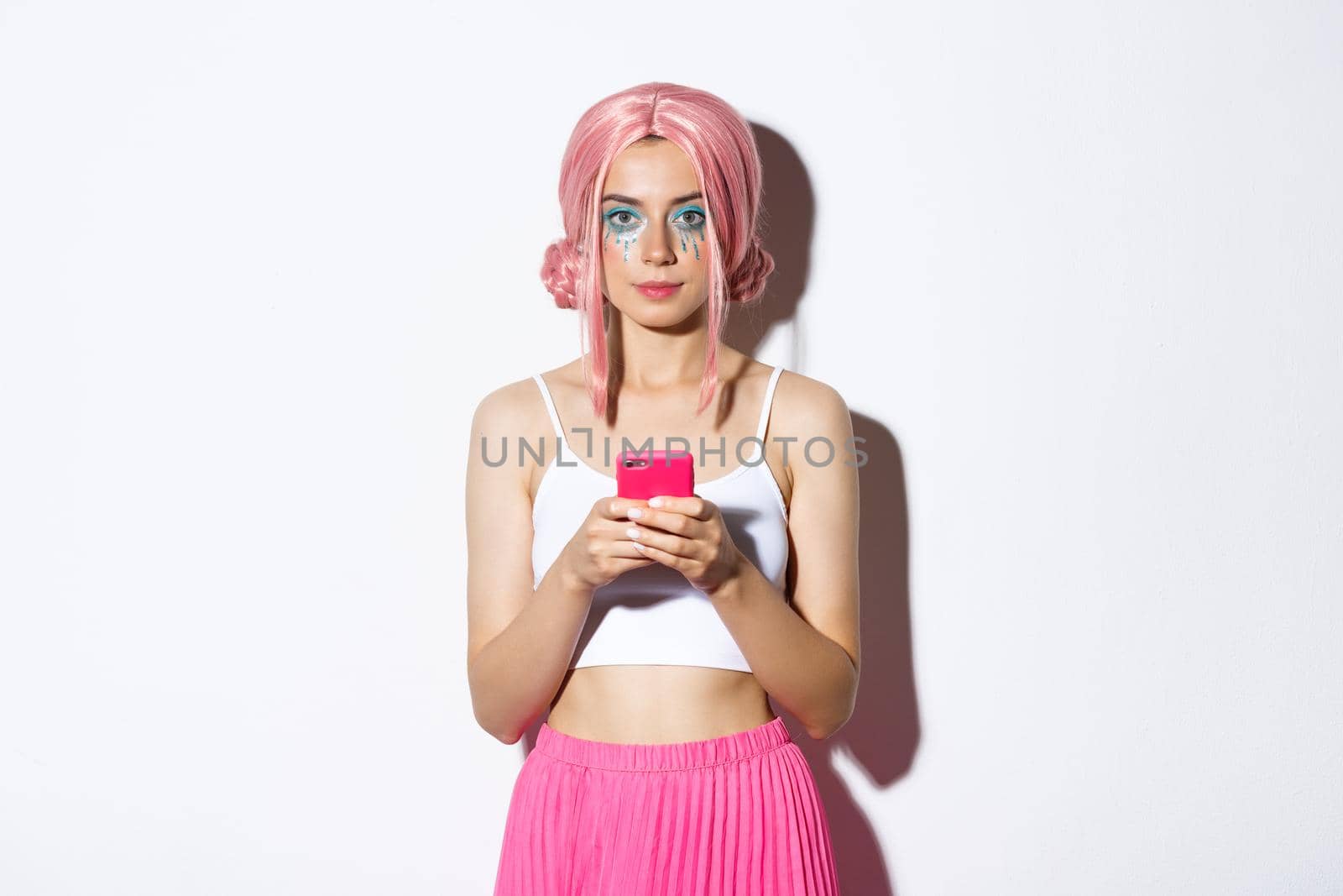 Portrait of attractive party girl in pink wig, with bright makeup, holding smartphone and looking at camera confident by Benzoix