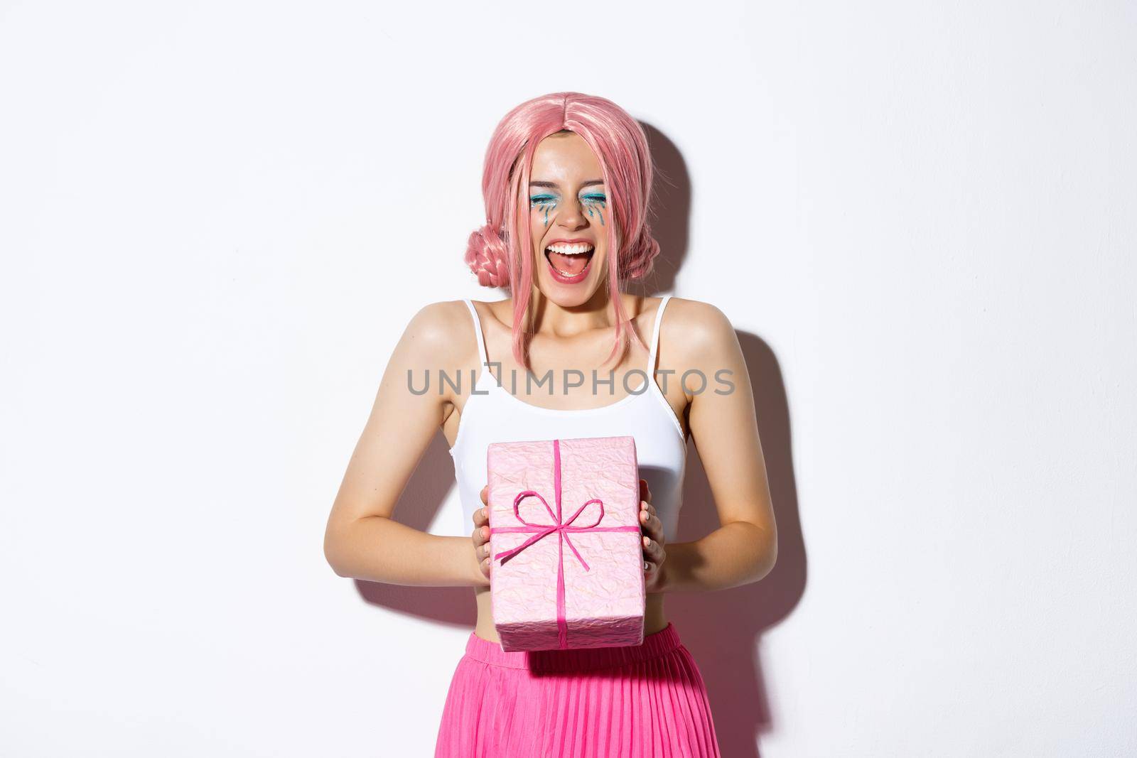 Cheerful birthday girl looking excited, wearing pink wig, shouting of joy, receiving bday gift, standing over white background and celebrating.