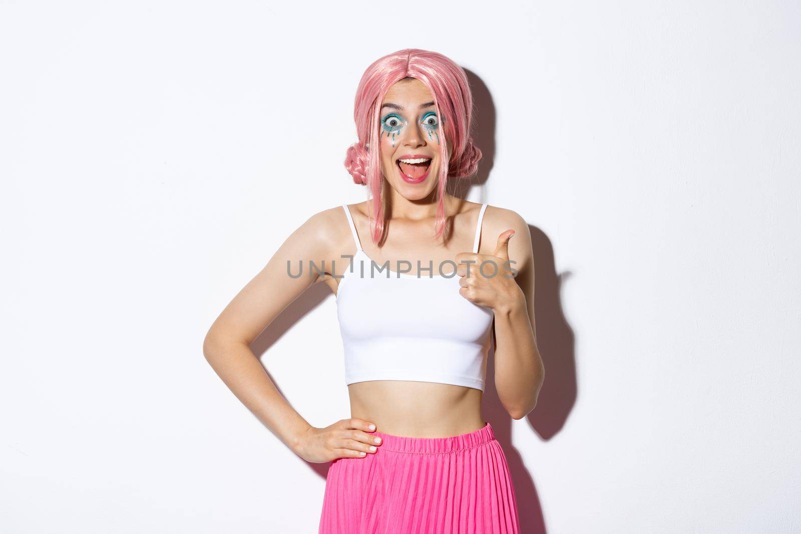 Portrait of enthusiastic attractive female model with pink hair and party makeup, showing thumbs-up in approval, smiling amazed, standing over white background in outfit for halloween.