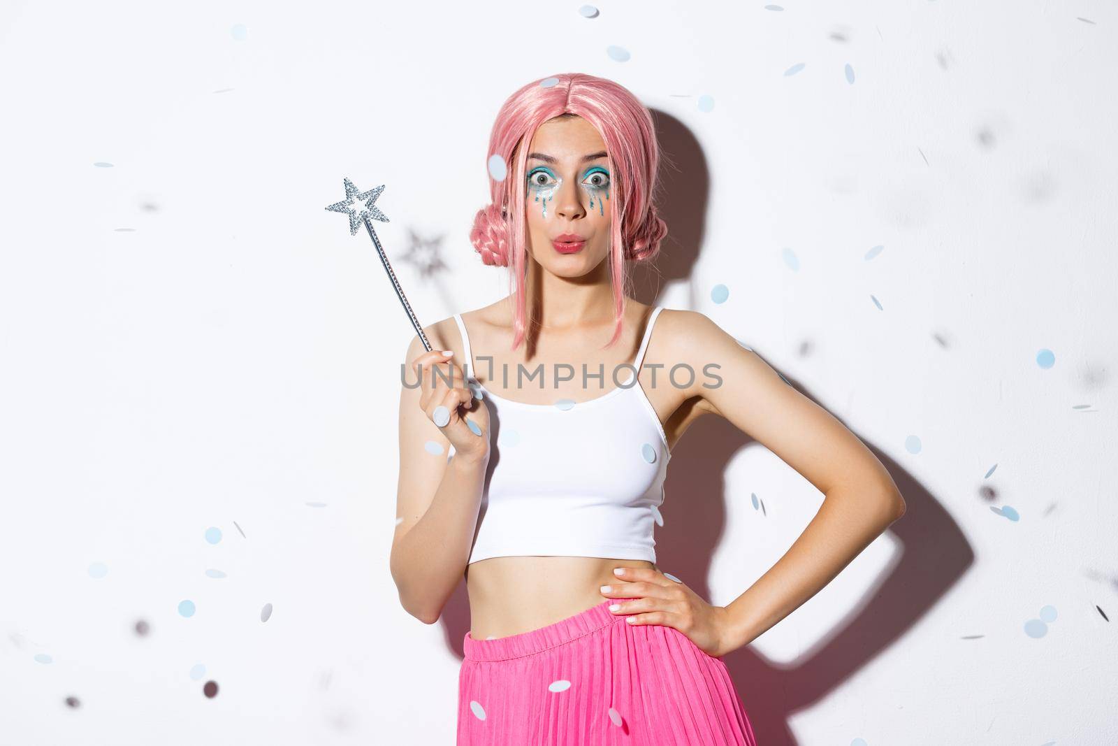 Attractive girl with magic wand celebrating halloween in fairy costume and pink wig, looking coquettish at camera, standing over white background while confetti falling down.