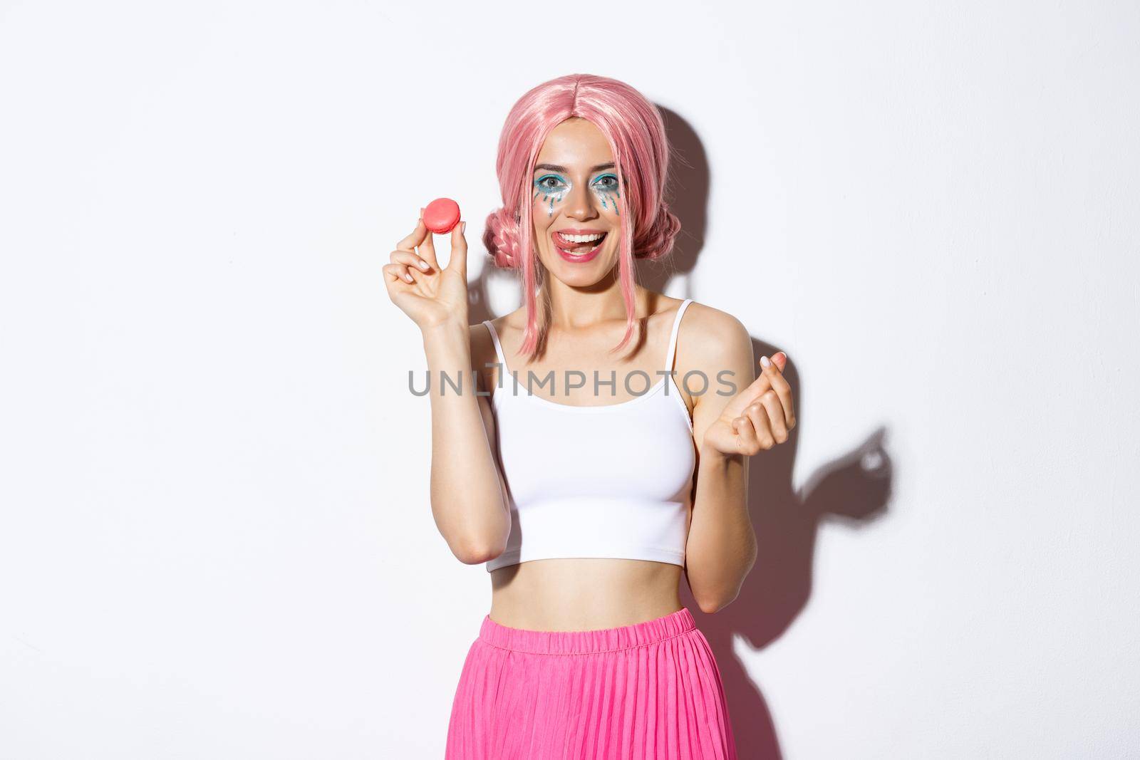 Image of beautiful girl with bright makeup eating tasty macaroons, wearing pink wig, showing tongue and smiling, standing over white background by Benzoix