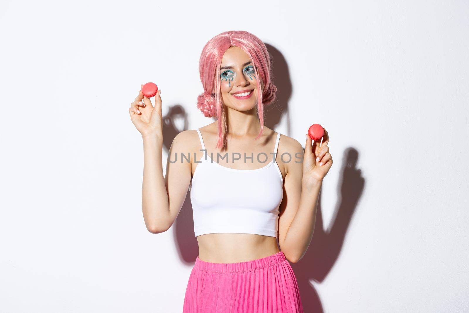 Lovely young woman in pink wig showing macaroons and smiling, eating sweets, standing over white background by Benzoix