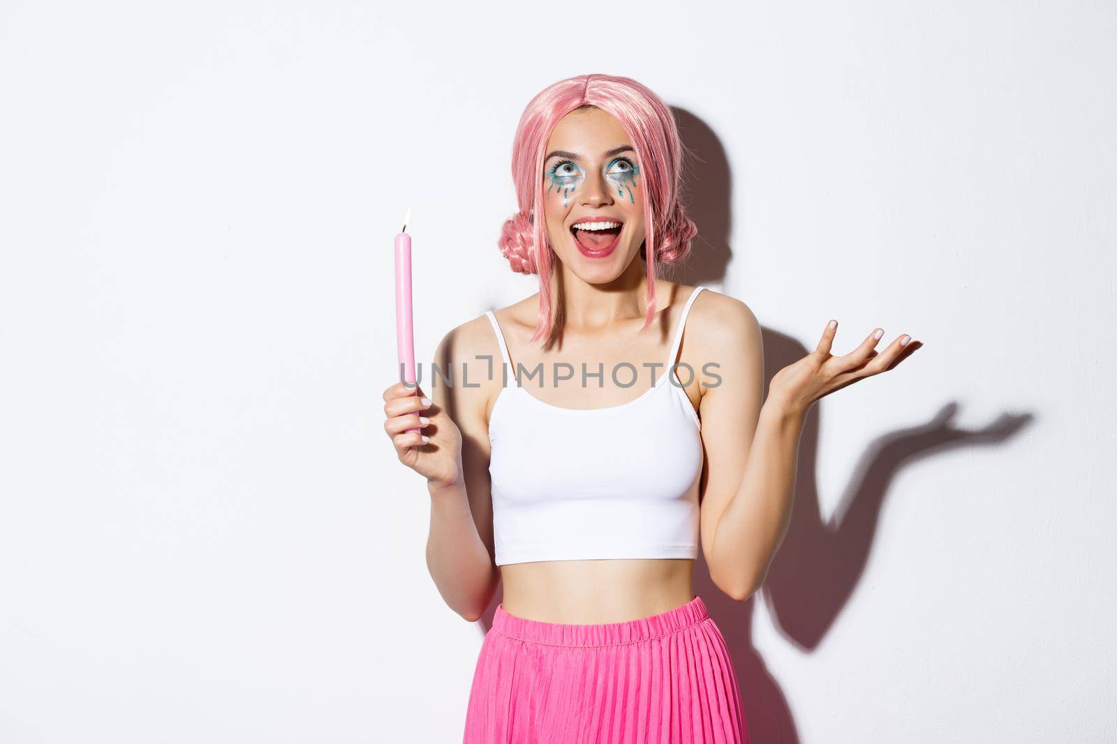 Portrait of excited beautiful girl dressed up as fairy on halloween, wearing pink wig and bright makeup, holding candle and talking to someone, standing over white background.