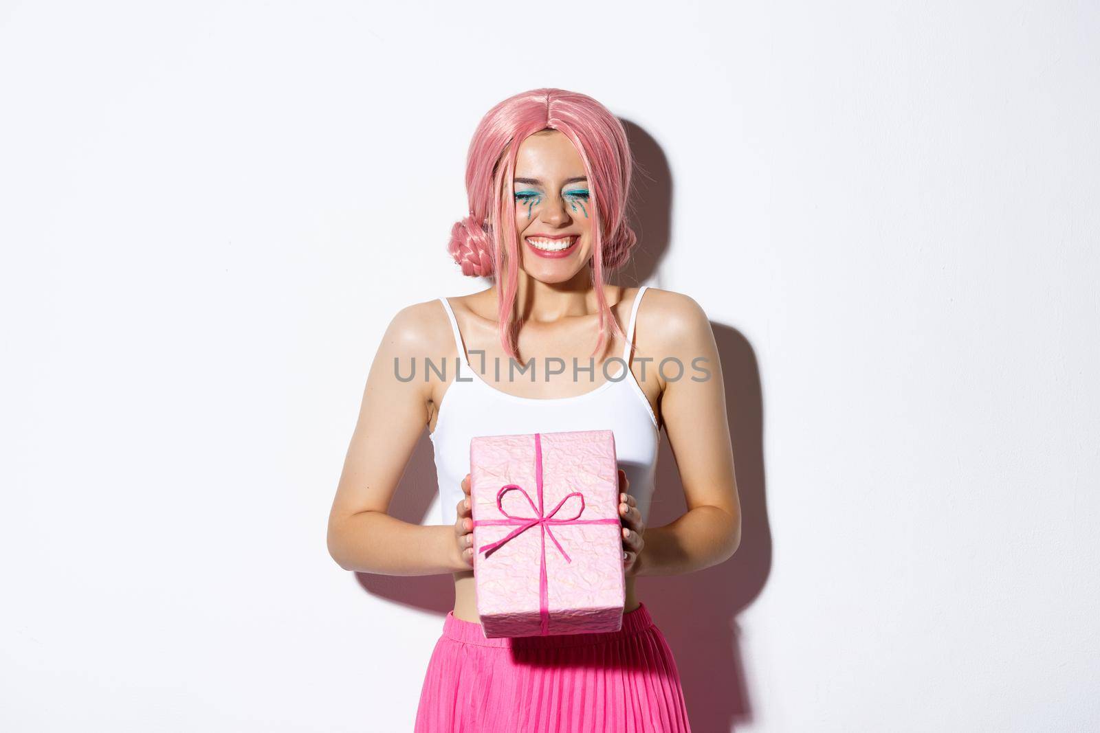 Portrait of happy beautiful b-day girl looking excited, receiving birthday gift and smiling joyfully, standing in pink wig and party outfit over white background.