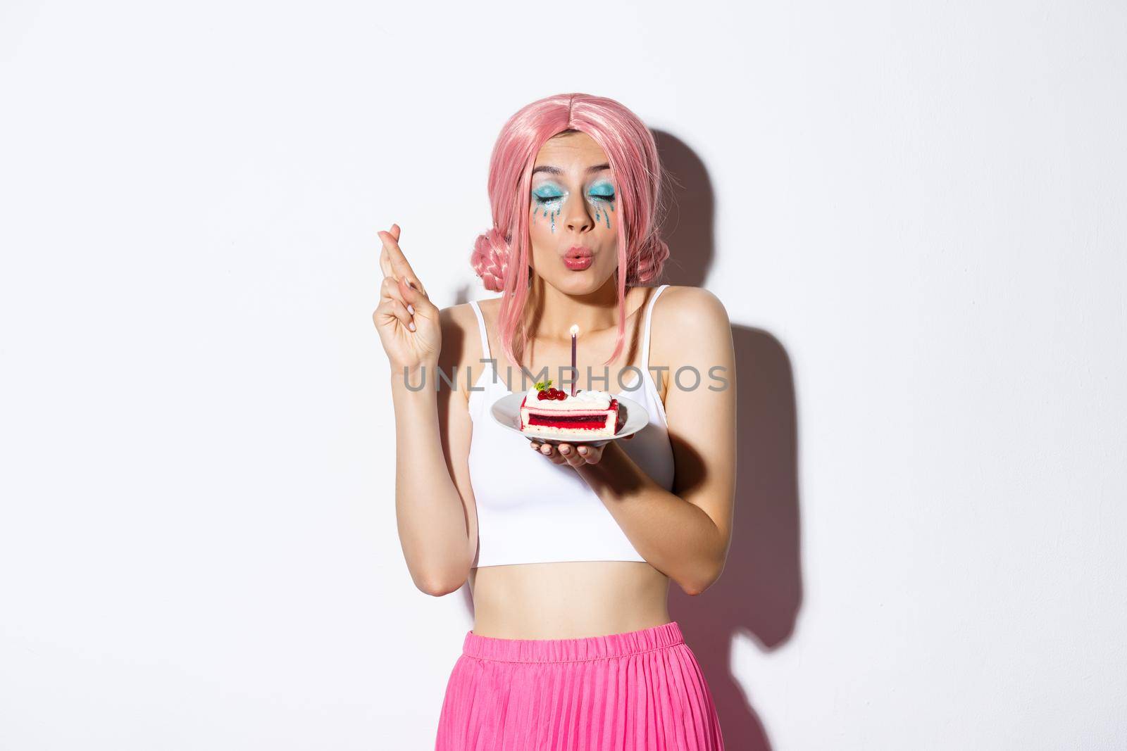 Portrait of hopeful birthday girl in pink wig, making wish with fingers crossed, holding b-day cake, standing over white background.