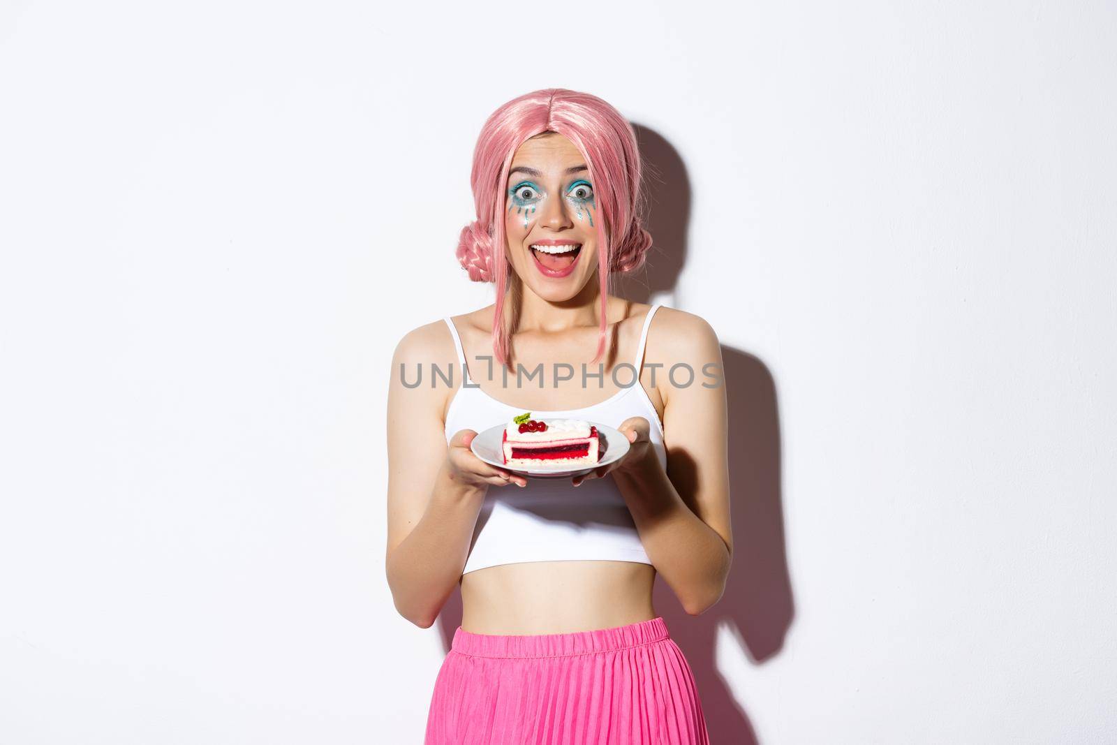 Portrait of excited birthday girl celebrating, wearing pink wig, holding b-day cake and smiling happy, standing over white background.