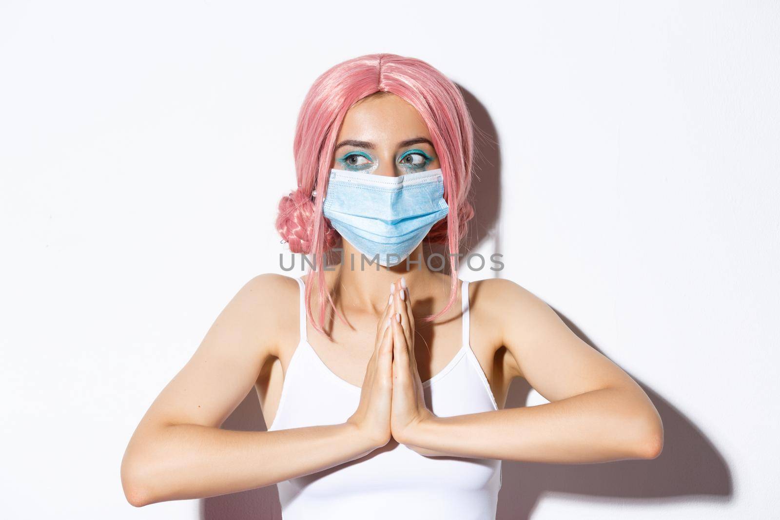 Covid-19, social distancing and people concept. Close-up of cute party girl in pink wig and medical mask, holding hands together and looking left, begging for help by Benzoix