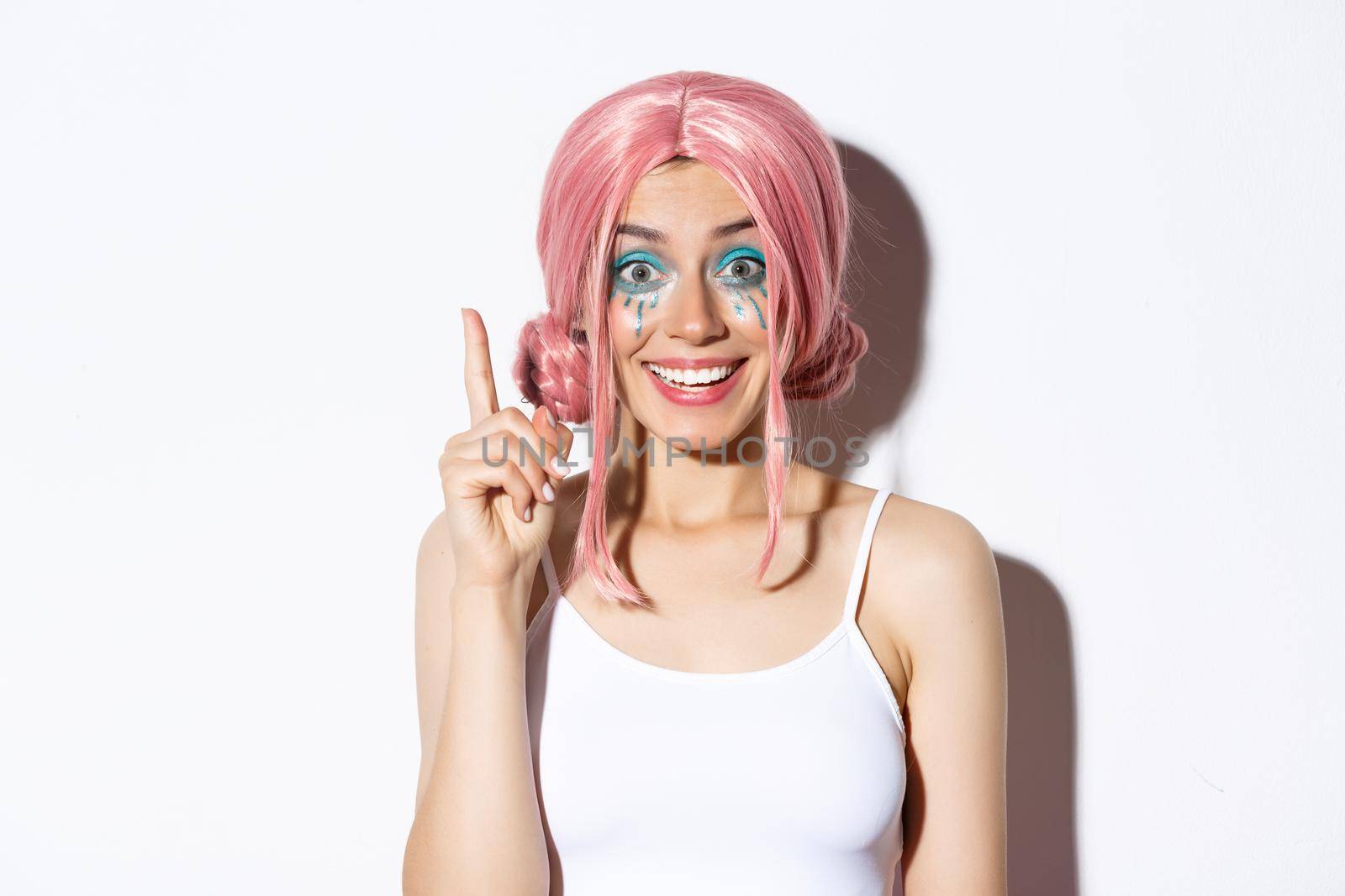 Close-up of woman in pink party wig and bright makeup, have idea, raising index finger in eureka sign, standing over white background by Benzoix