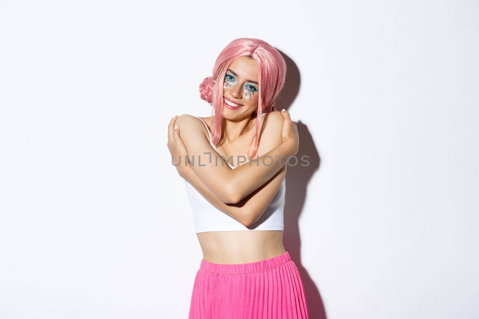 Portrait of beautiful and tender young woman celebrating halloween in glamorous outfit with pink wig, embracing her body and smiling at camera, standing over white background.