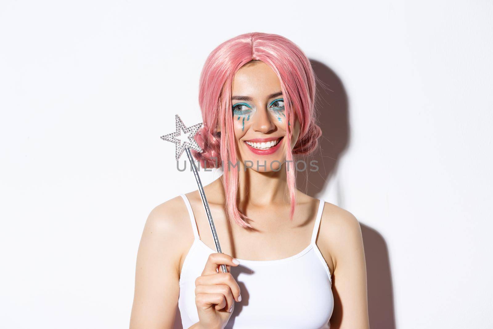 Close-up of lovely beautiful girl in halloween costume of fairy, holding magic wand and smiling, looking left at your logo, standing over white background.