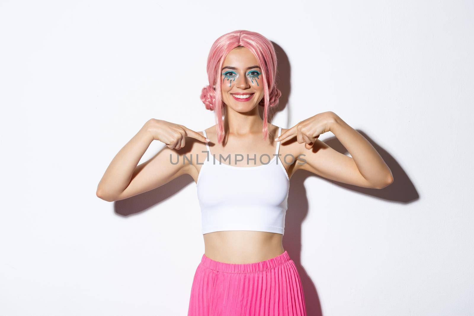 Portrait of cheerful party girl with pink wig and bright makeup, pointing at herself and smiling confident, showing her halloween outfit, standing over white background by Benzoix