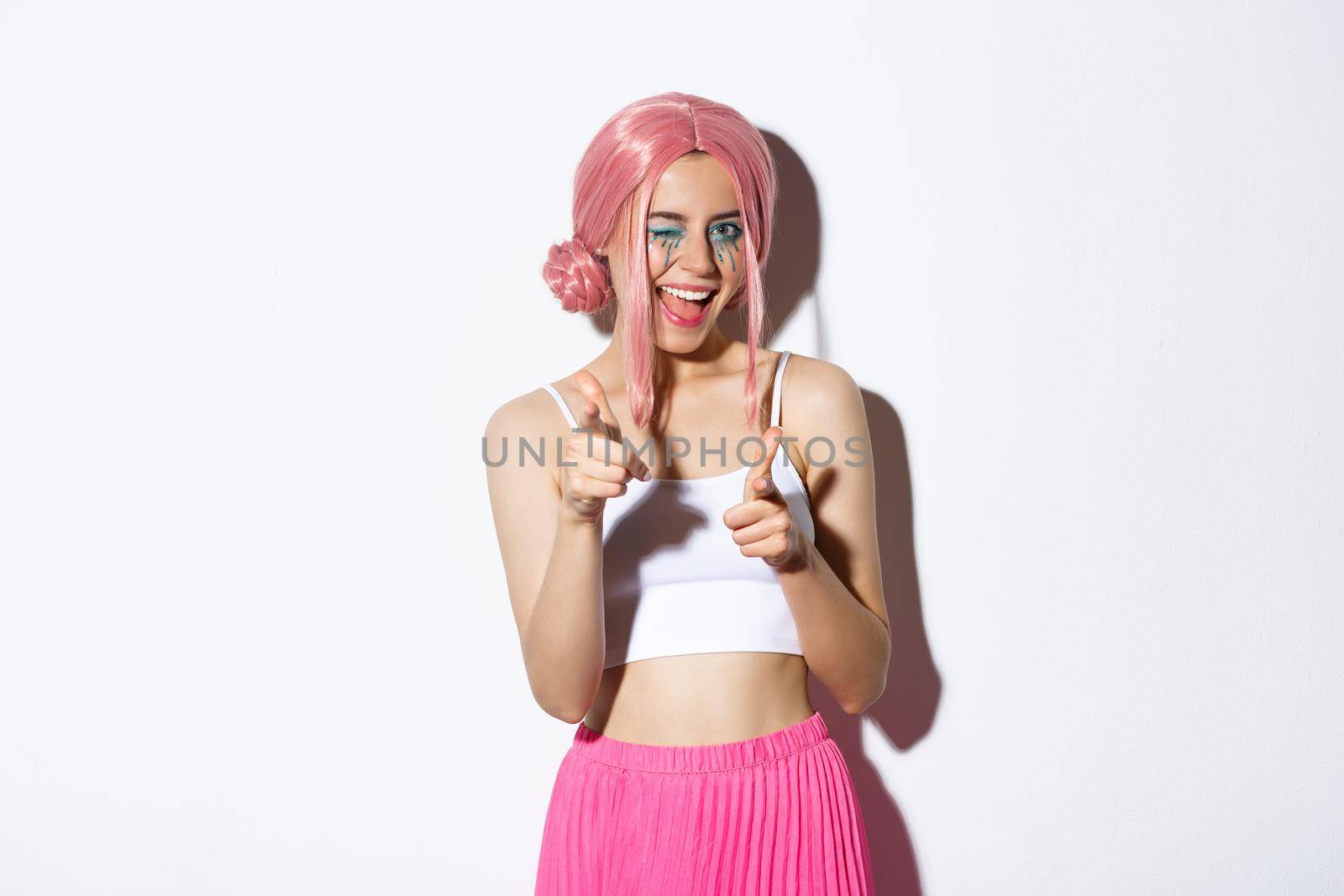 Sassy attractive girl in halloween costume and pink wig, pointing at camera and smiling, congratulating or praising someone, standing over white background.