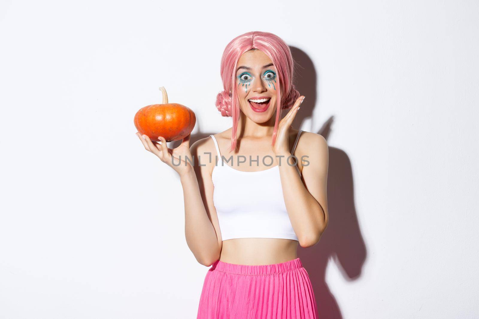 Portrait of surprised girl in pink wig, holding pumpkin and looking excited, celebrating halloween, standing over white background.