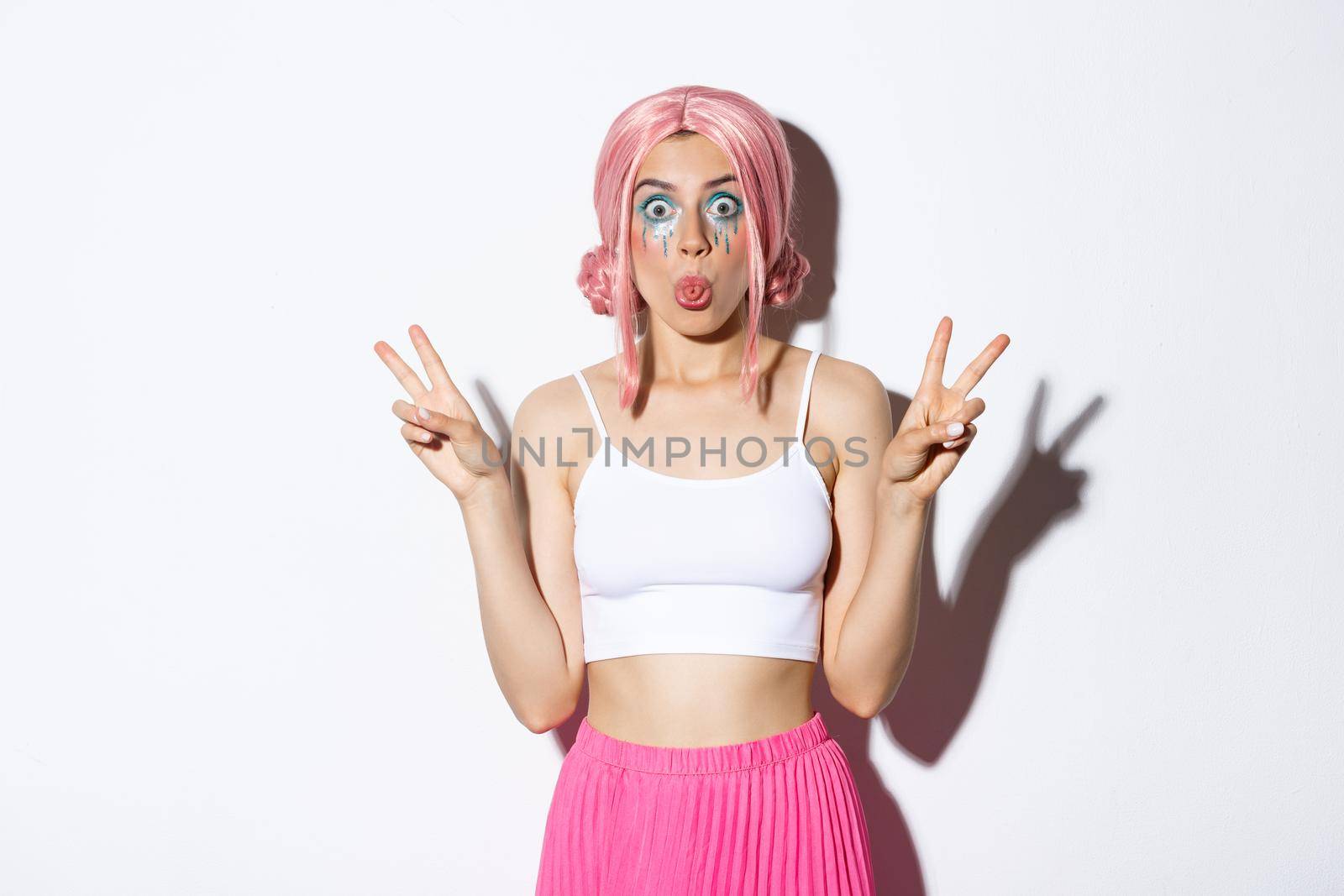 Image of silly girl in pink wig, celebrating halloween, showing funny faces and sticking tongue, make peace signs, standing over white background.